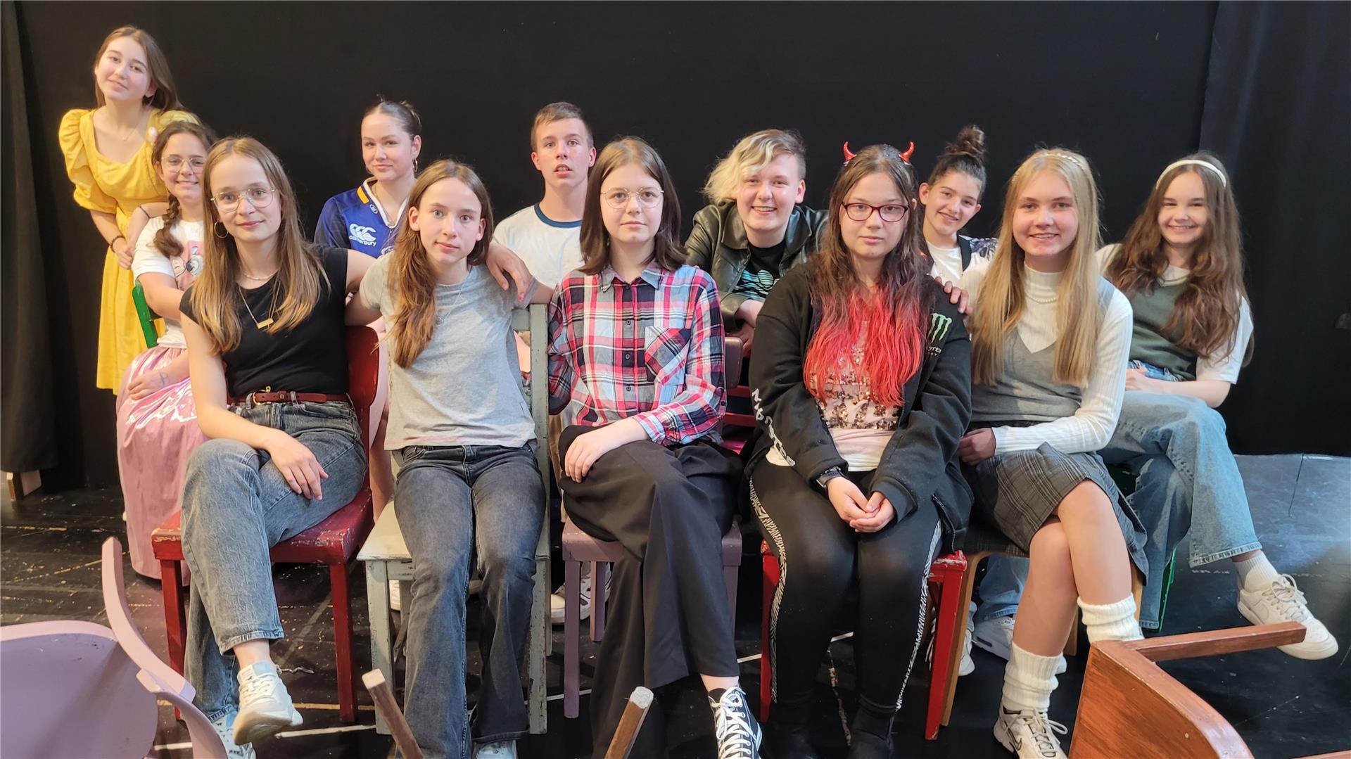 Gruppenfoto von Jugendlichen des Theaterlabors 3 des Stadttheaters Bremerhaven, sitzend und stehend in Kostümen und Alltagskleidung vor schwarzem Bühnenhintergrund.