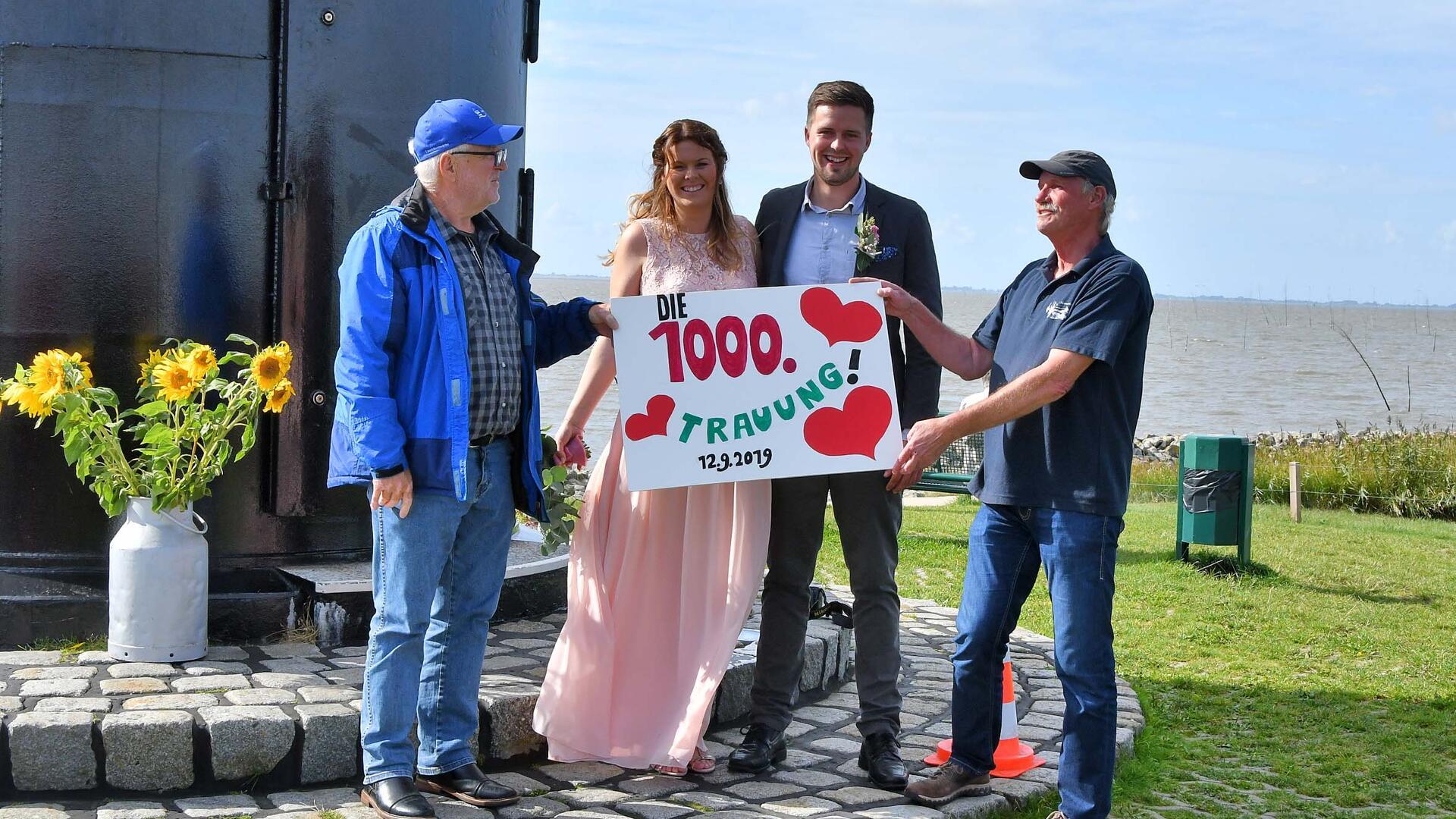 Vor einem schwarzen Leuchtturm an der Küste stehen ein Brautpaar und zwei Männer. Alle lächeln und halten gemeinsam ein Schild mit der Aufschrift „Die 1000. Trauung! 12.9.2019“, verziert mit roten Herzen. Links steht eine Vase mit Sonnenblumen.