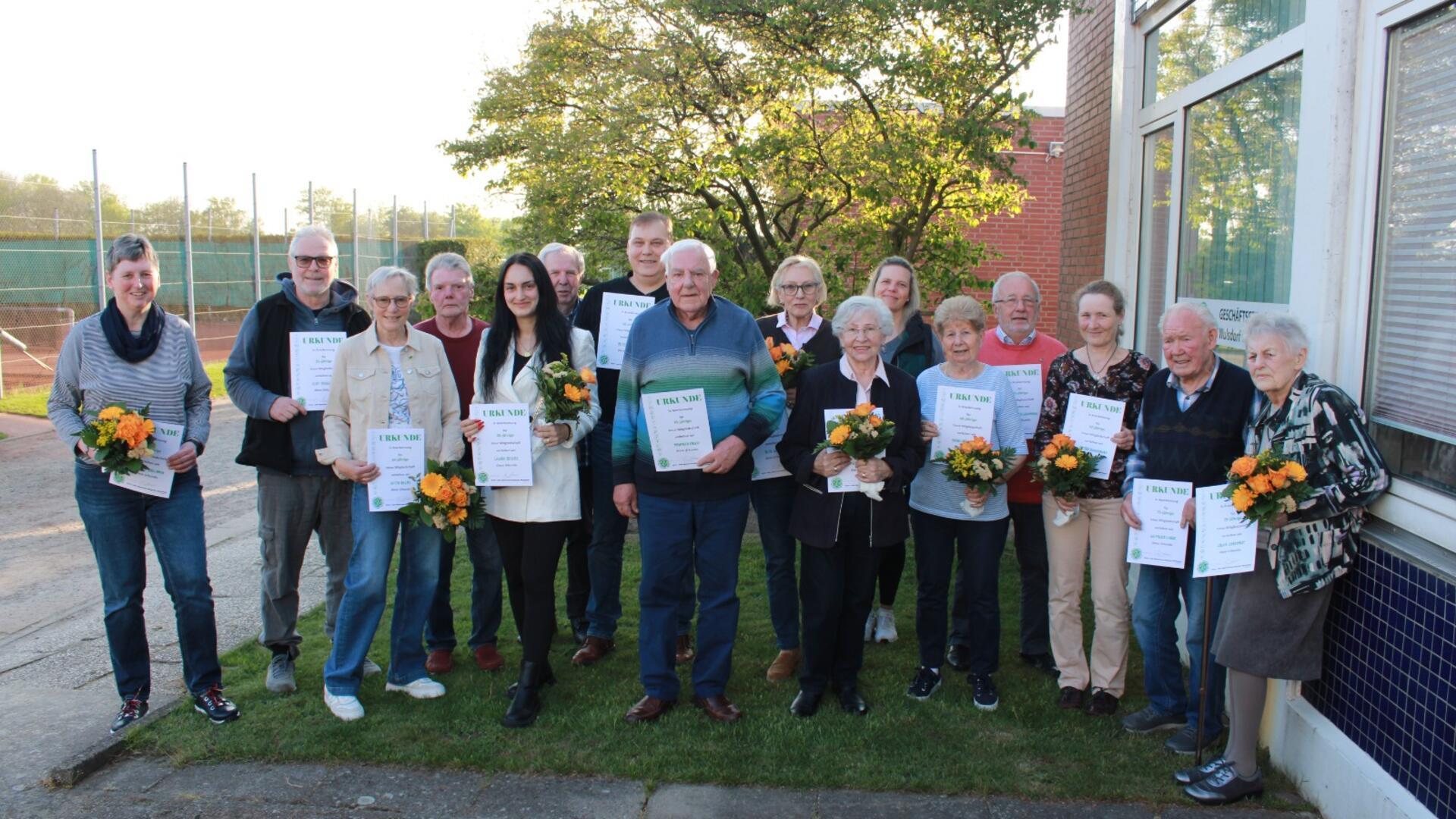 16 geehrte Mitglieder der TSV Wulsdorf halten Urkunden und Blumensträuße in den Händen und lächeln in die Kamera.