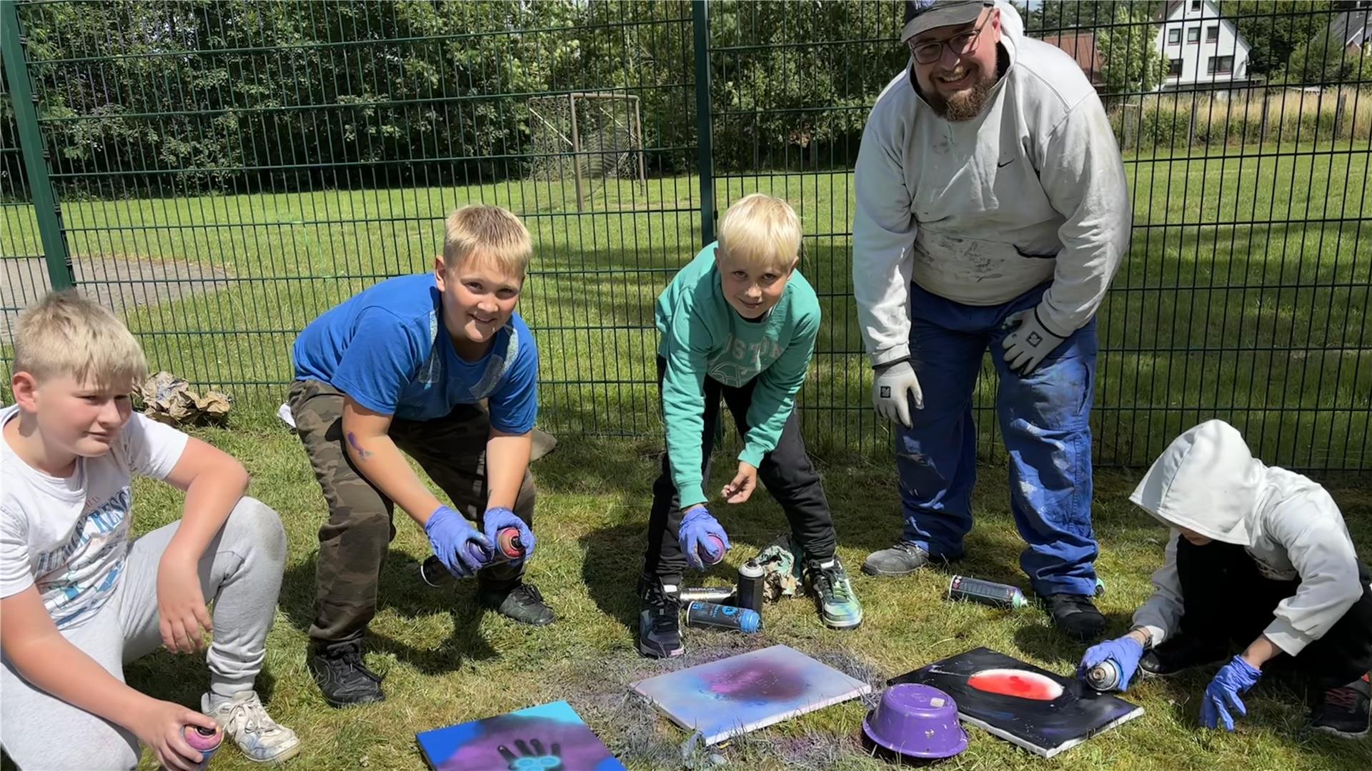 Kinder und ein Betreuer gestalten im Freien kreative Bilder mit Farbspraydosen; sie arbeiten auf dem Rasen vor einem Zaun, umgeben von buntem Material.