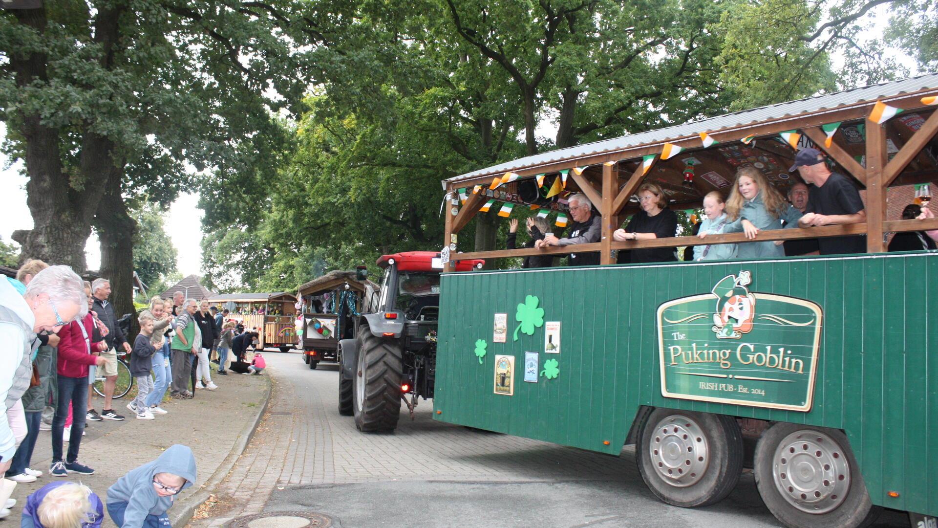 Traktoren ziehen Festwagen über eine Dorfstraße. Am Rand stehen Menschen. 