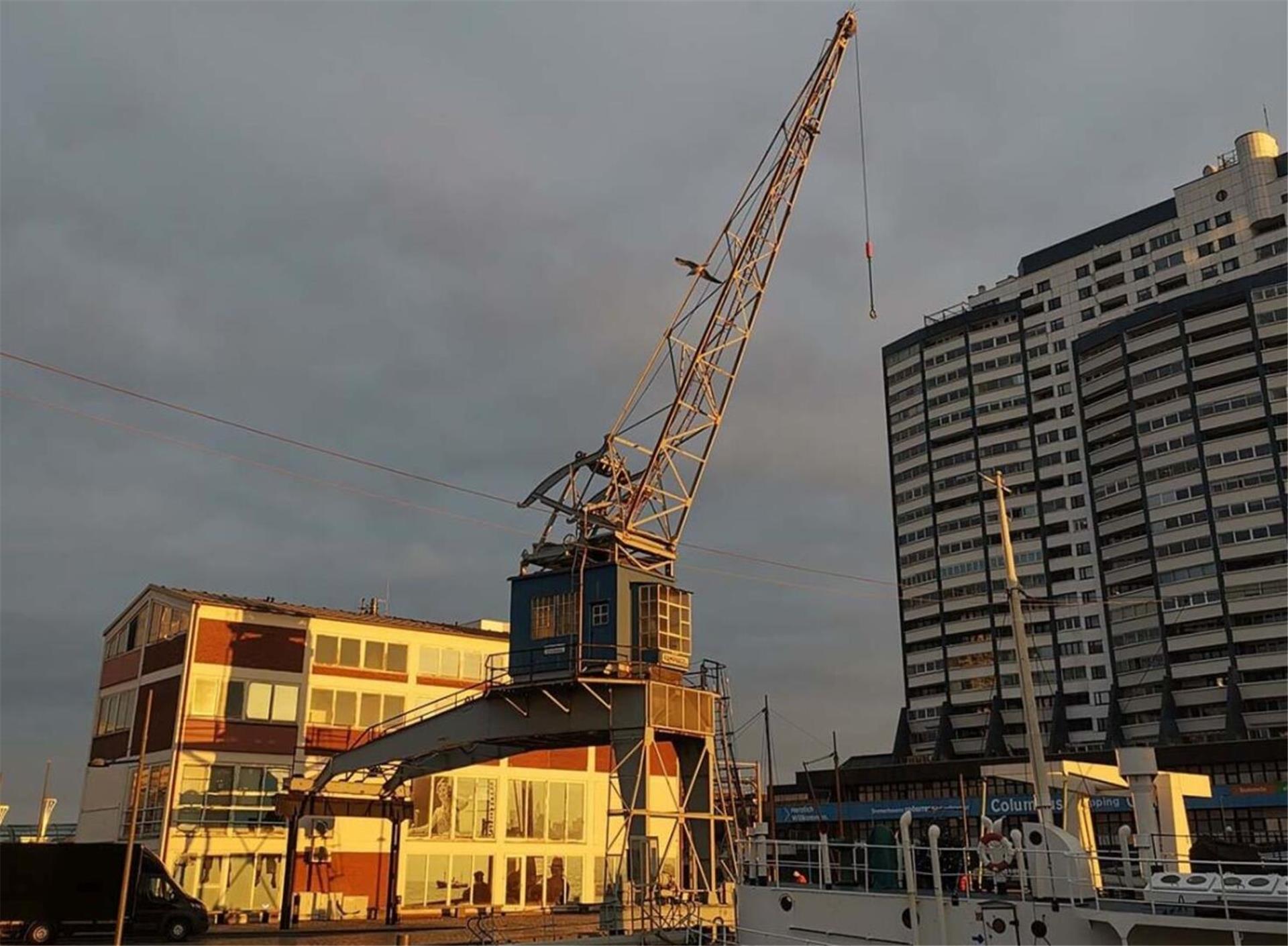 Historischer Hafenkran bei Sonnenuntergang in Bremerhaven, im Vordergrund ist ein Teil eines weißen Schiffs zu sehen, im Hintergrund ein modernes Hochhaus und ein Backsteingebäude mit großen Fenstern.