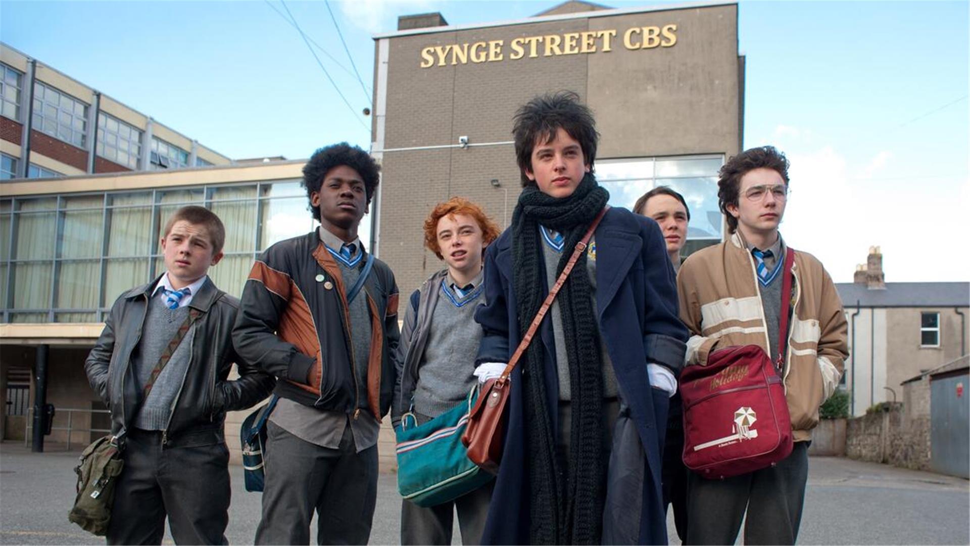 Sechs Jugendliche stehen vor einem Gebäude, an dem der Schriftzug "Synge Street CBS" zu sehen ist. 80er-Hits und Jugendträume flimmern über die Leinwand: „Sing Street“ erzählt von Selbstfindung, erster Liebe und Freiheit - zu sehen am Sonnabend auf der Sommerstraße „Alte Bürger“ während der Reihe „Nachtflimmern – Filme in der Stadt“.