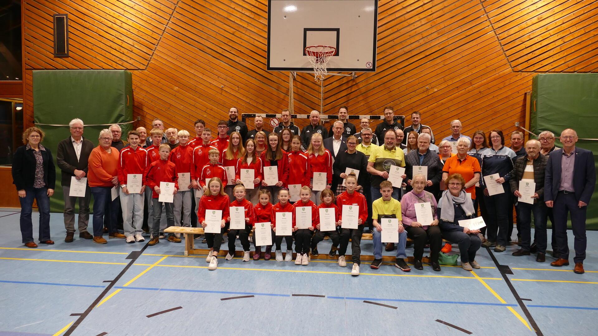 Eine große Gruppe von Menschen, darunter Kinder, Jugendliche und Erwachsene verschiedener Altersgruppen, posiert in einer Sporthalle für ein Gruppenfoto. Viele der Personen tragen rote Sportbekleidung, während andere in Alltagskleidung oder Vereinskleidung gekleidet sind. Sie halten Zertifikate oder Urkunden in den Händen. 