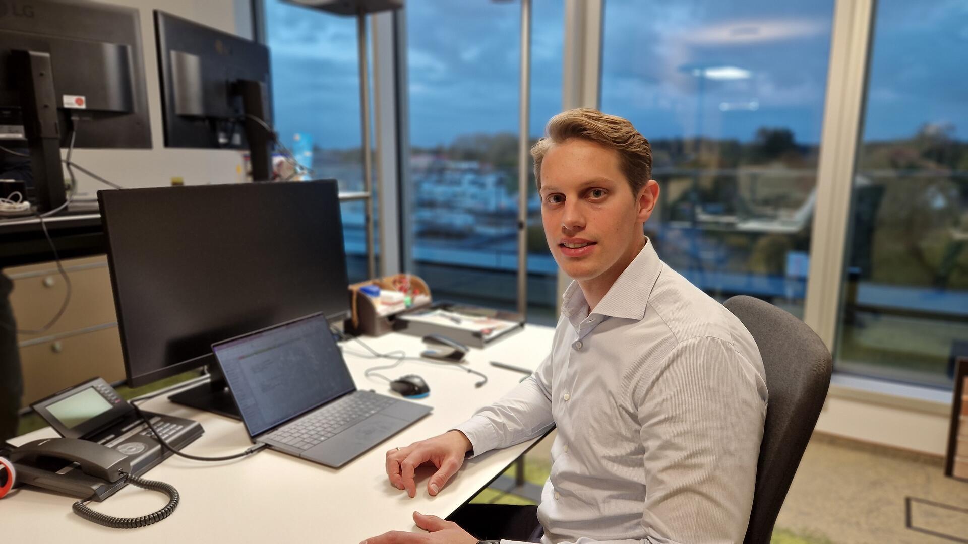 Ein Mann sitzt an einem Schreibtisch, auf dem ein PC steht, und lächelt in die Kamera.