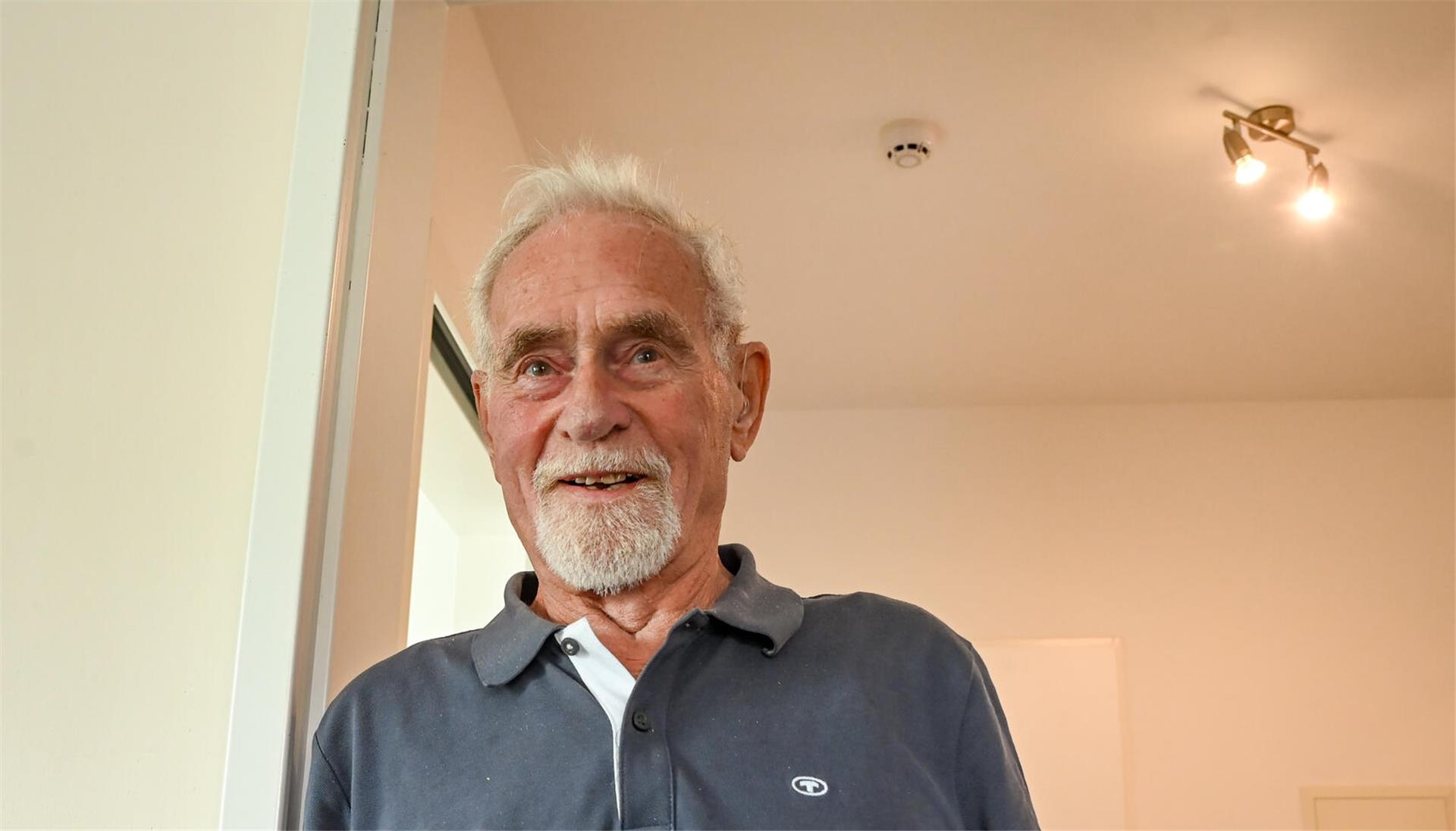 Ein Mann mit weißen Haaren und weißem Bart, der ein dunkles Polo-Shirt trägt. Er steht neben einer Tür, an der Decke hängt eine Lampe, daneben ist ein Rauchmelder zu sehen.