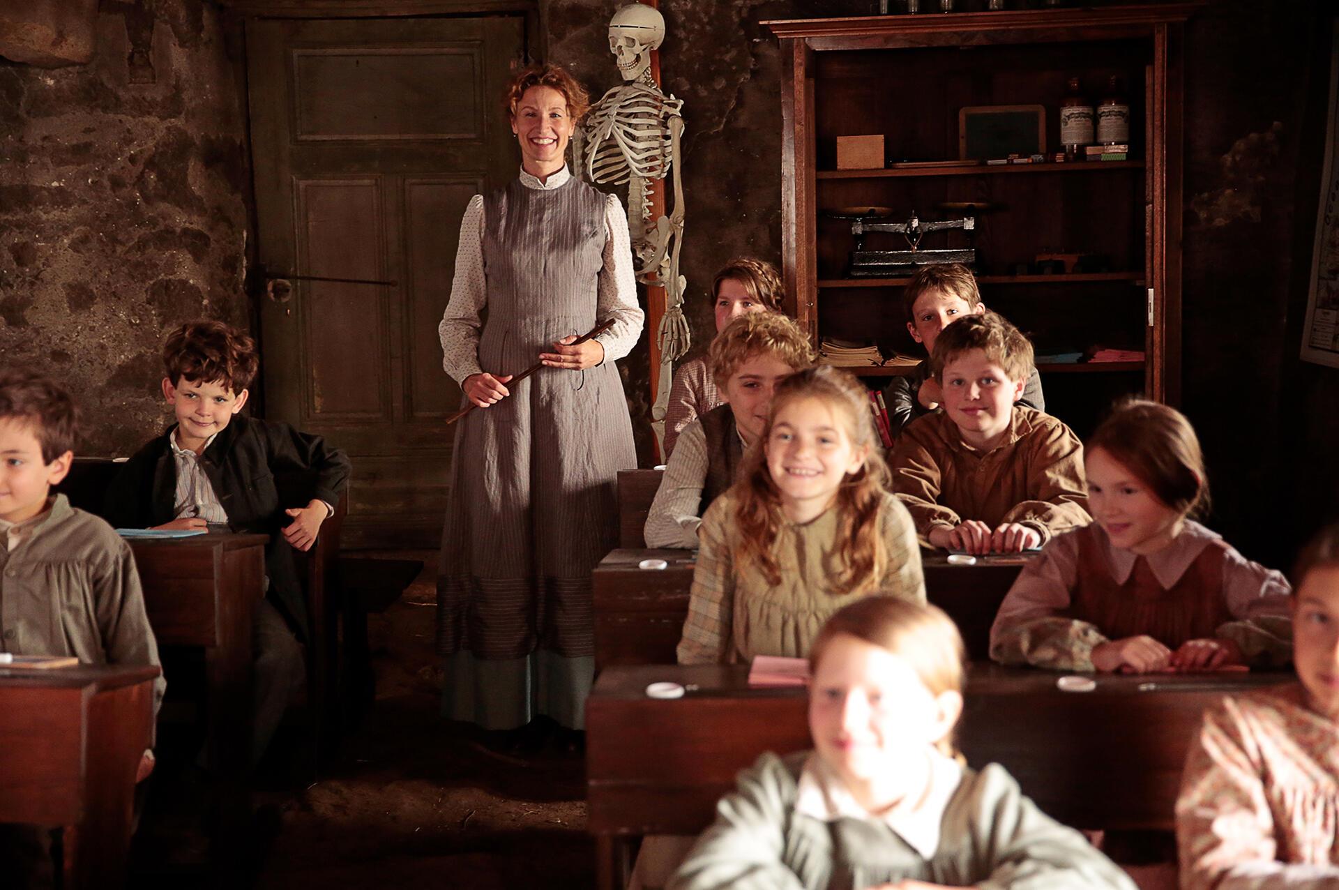 Eine lächelnde Lehrerin in einem Klassenzimmer mit mehreren Schülern. Alexandra Lamy (Mitte) als Louise Violet in einer Szene des Films „Louise und die Schule der Freiheit“.