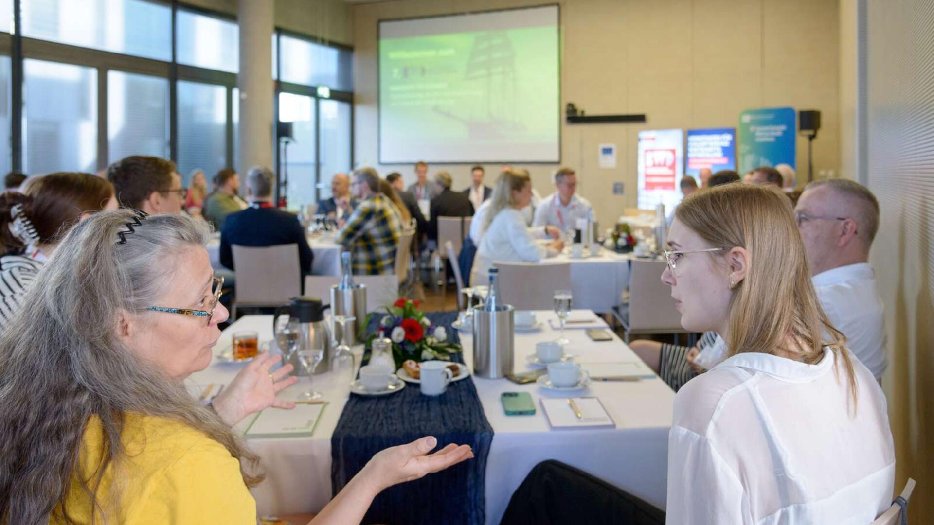 Menschen sitzen an Tischen und diskutieren, während im HIntergrund eine Leinwand zu sehen ist. 