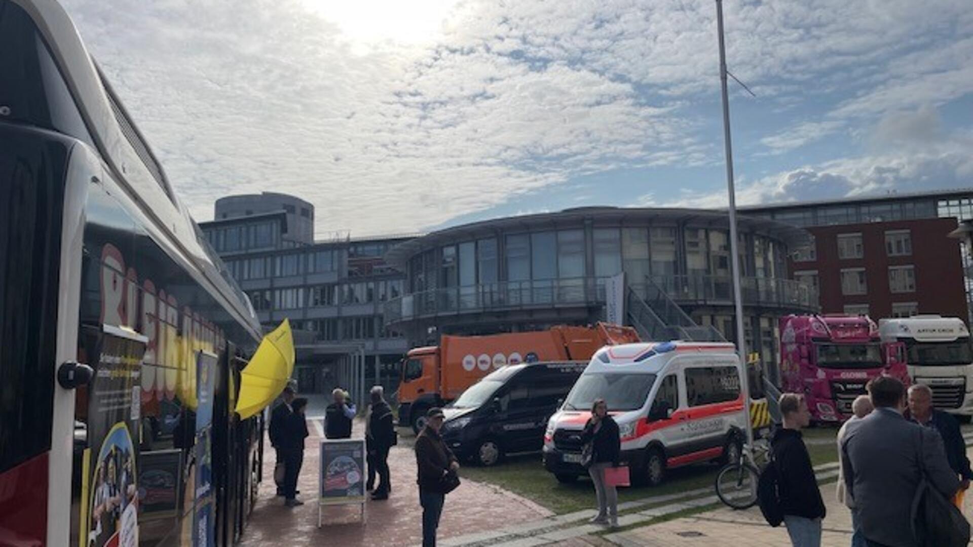 Ein Omnibus, ein Rettungswagen, ein Müllwagen und weitere Fahrzeuge stehen umringt von Menschen vor einem großen Bürogebäude. 
