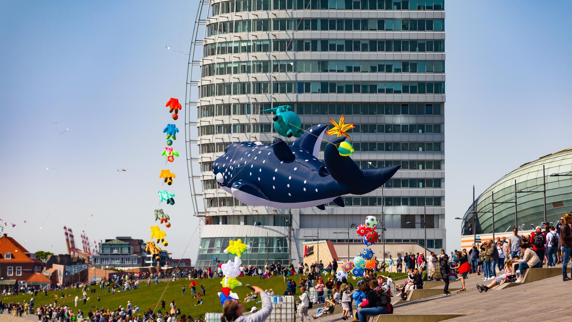 Menschen beobachten bunte Riesendrachen am Himmel beim Drachenfestival in Bremerhaven vor dem markanten Sail City Gebäude und den Havenwelten.