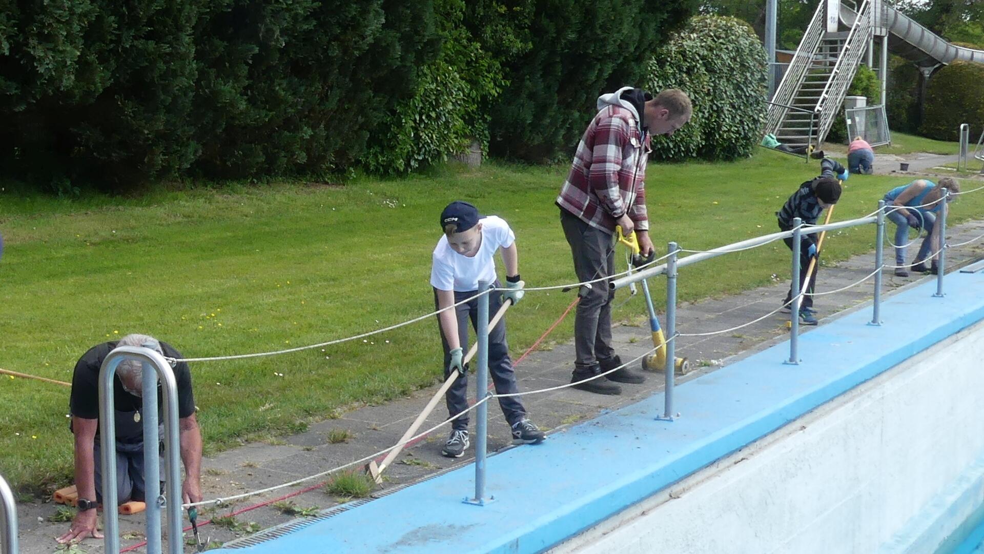 Mehrere Personen unterschiedlichen Alters arbeiten gemeinsam am Beckenrand eines leeren Freibads. Sie entfernen Unkraut und reinigen den Gehweg entlang des Schwimmbeckens. Im Hintergrund sind Rasenflächen, Büsche und eine Rutsche zu sehen.