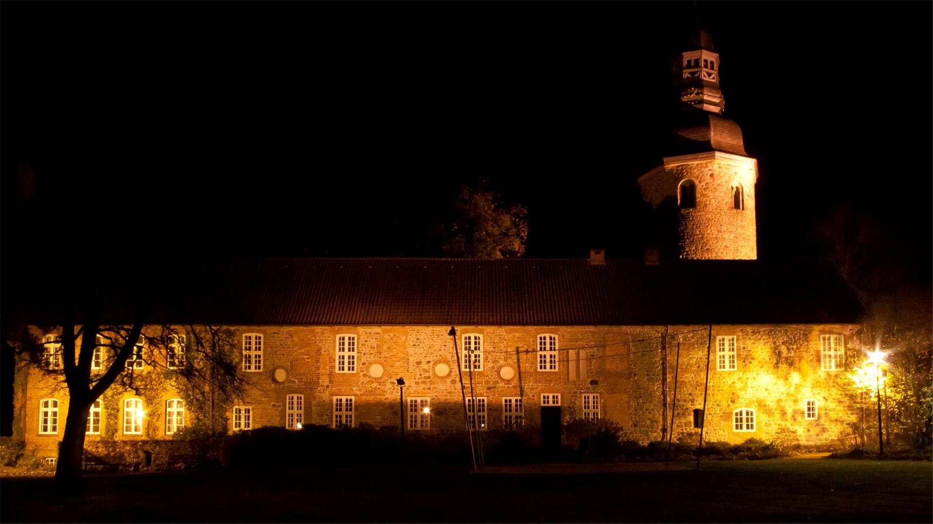 Ein langgestrecktes Gebäude und ein Kirchturm werden in der Dunkelheit angestrahlt.