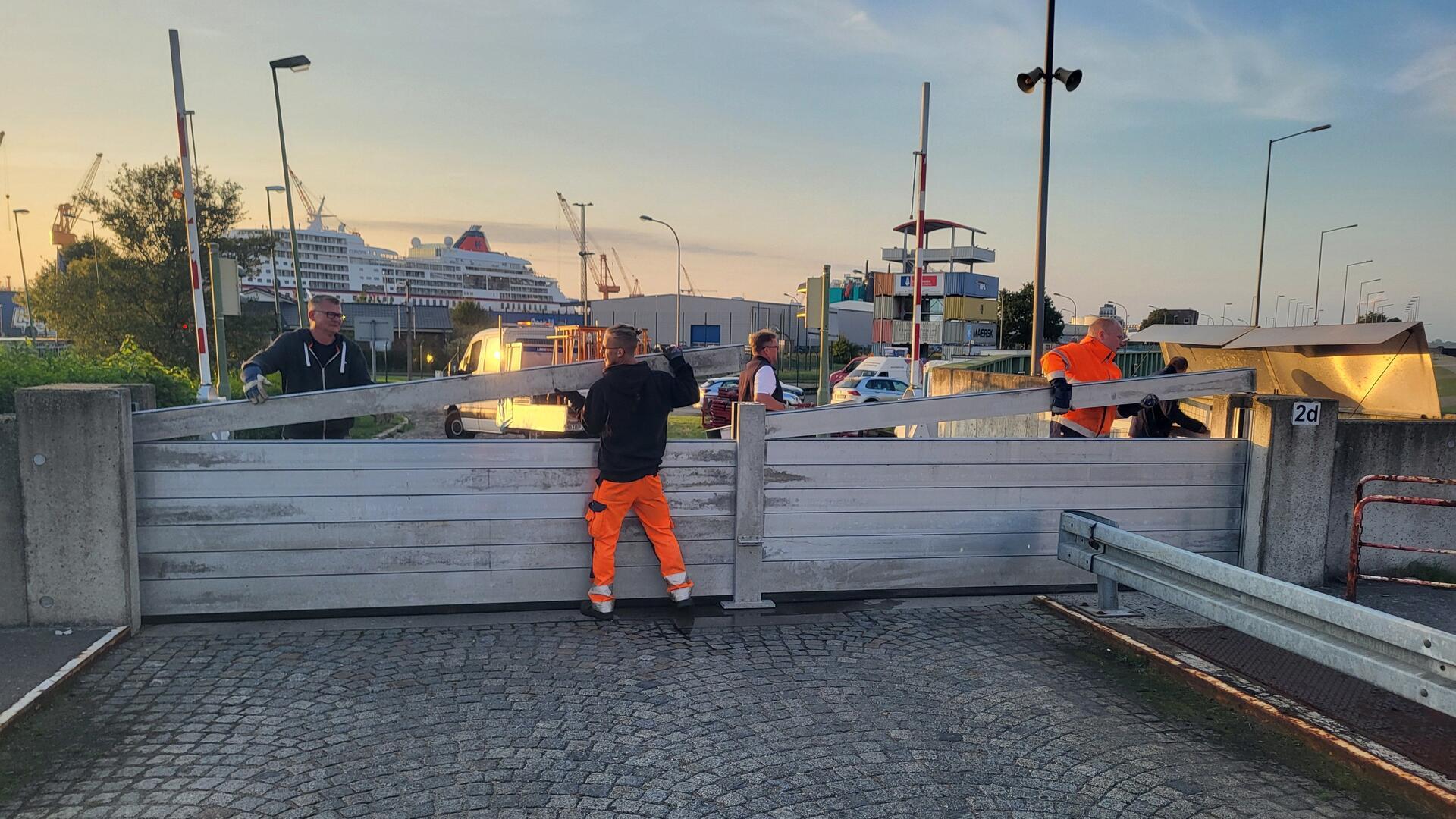 Einsatzkräfte bei der Montage eines Hochwasserschutztores. Im Hintergrund sind ein Kreuzfahrtschiff, Hafenanlagen und ein Aussichtsturm aus Containern zu sehen.