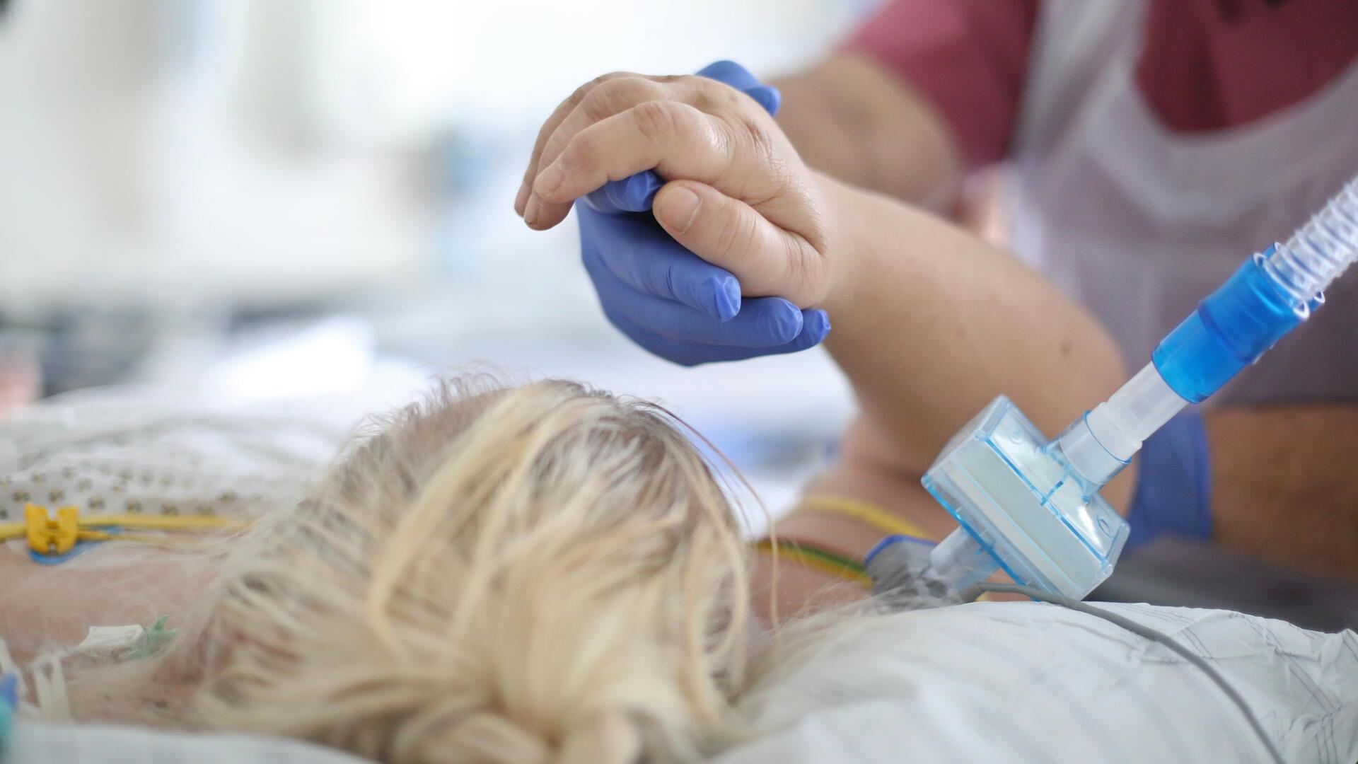 Pflegekraft mit blauen Handschuhen hält die Hand einer beatmeten Patientin auf der Intensivstation.