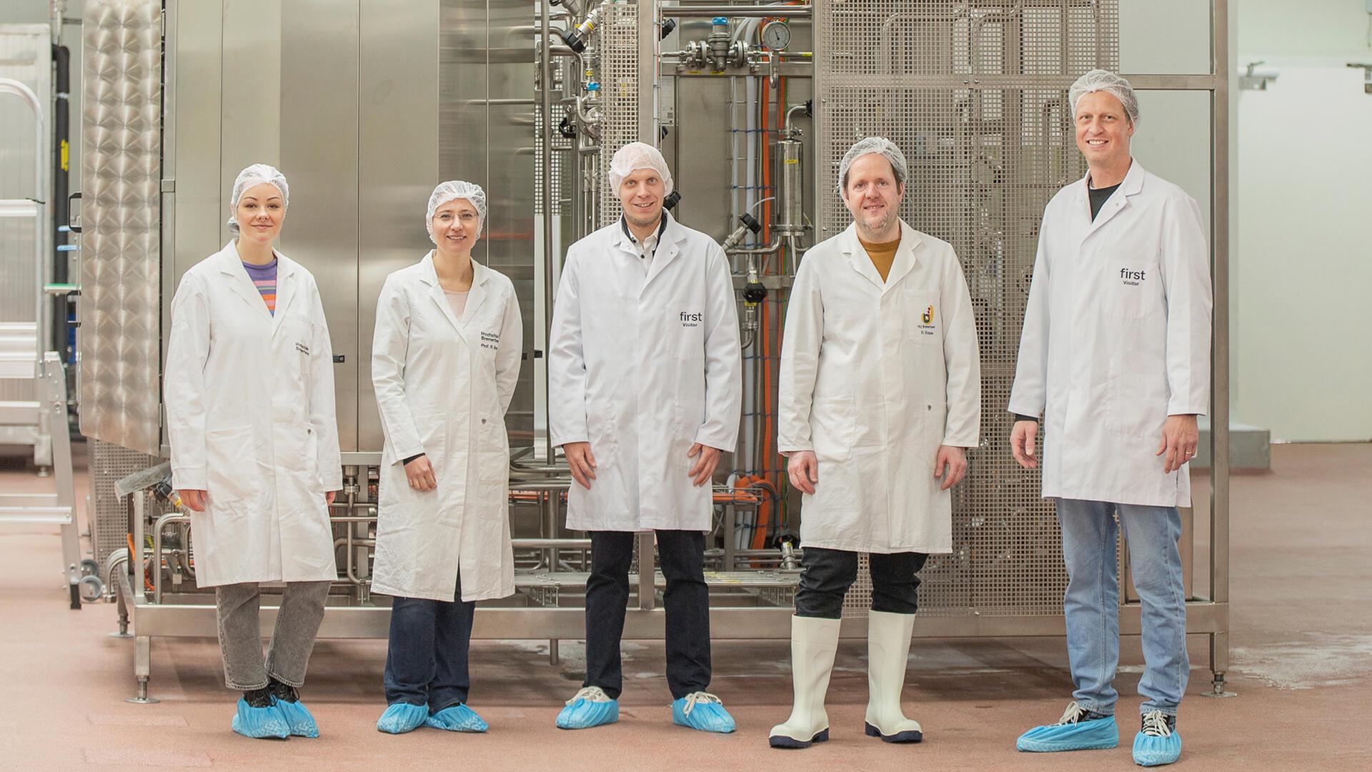 Zwei Frauen und drei Männer mit Hauben und weißen Kitteln stehen vor einer industriellen Anlage.