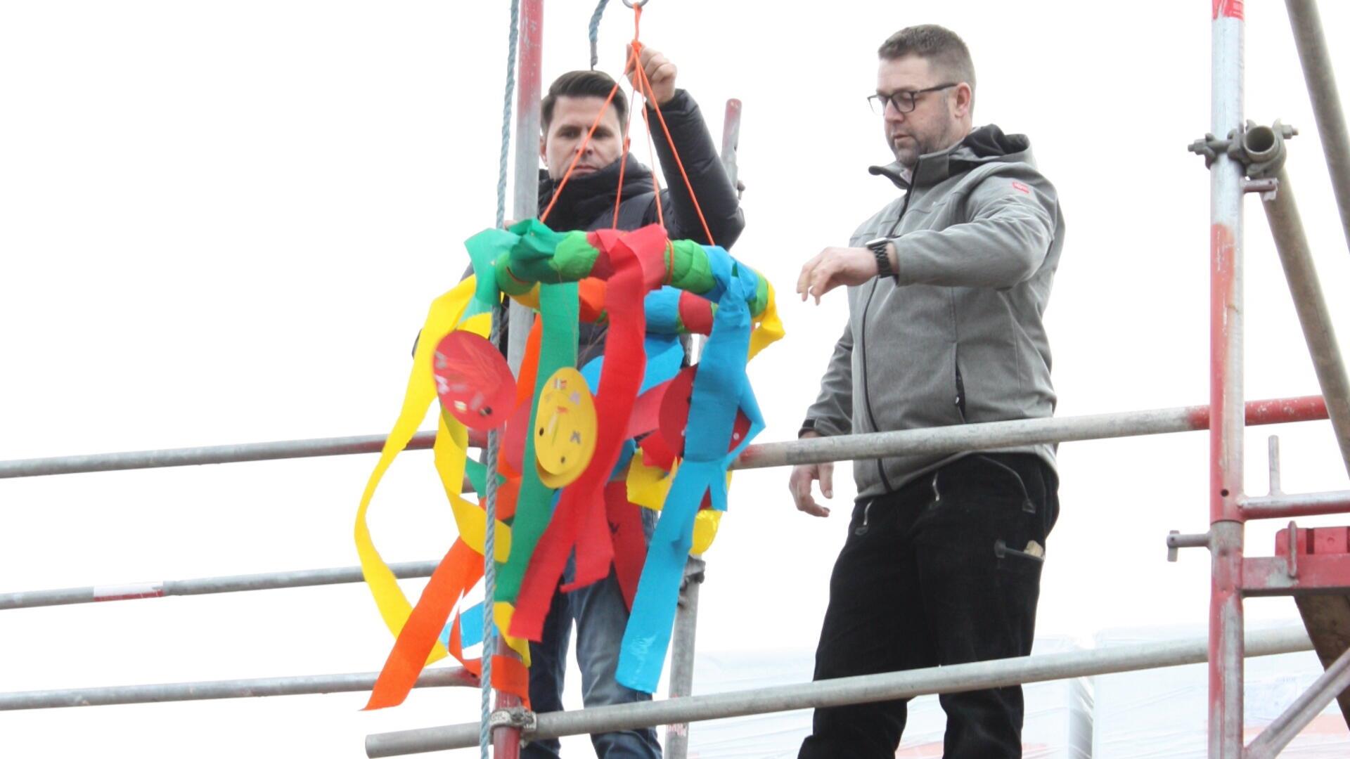 Architekt André (links) und Bauamt-Mitarbeiter Oliver Hünken (rechts) hängen gemeinsam die von der Kita "Rasselbande" gemachte Richtkrone auf.
