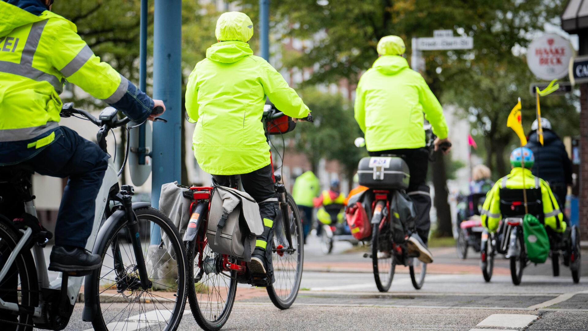 Mehrere Radfahrende in leuchtend gelben Warnjacken fahren in einer Gruppe durch eine Stadt. Auch ein Polizei-Radfahrer und ein Liegerad mit Wimpel sind zu sehen.