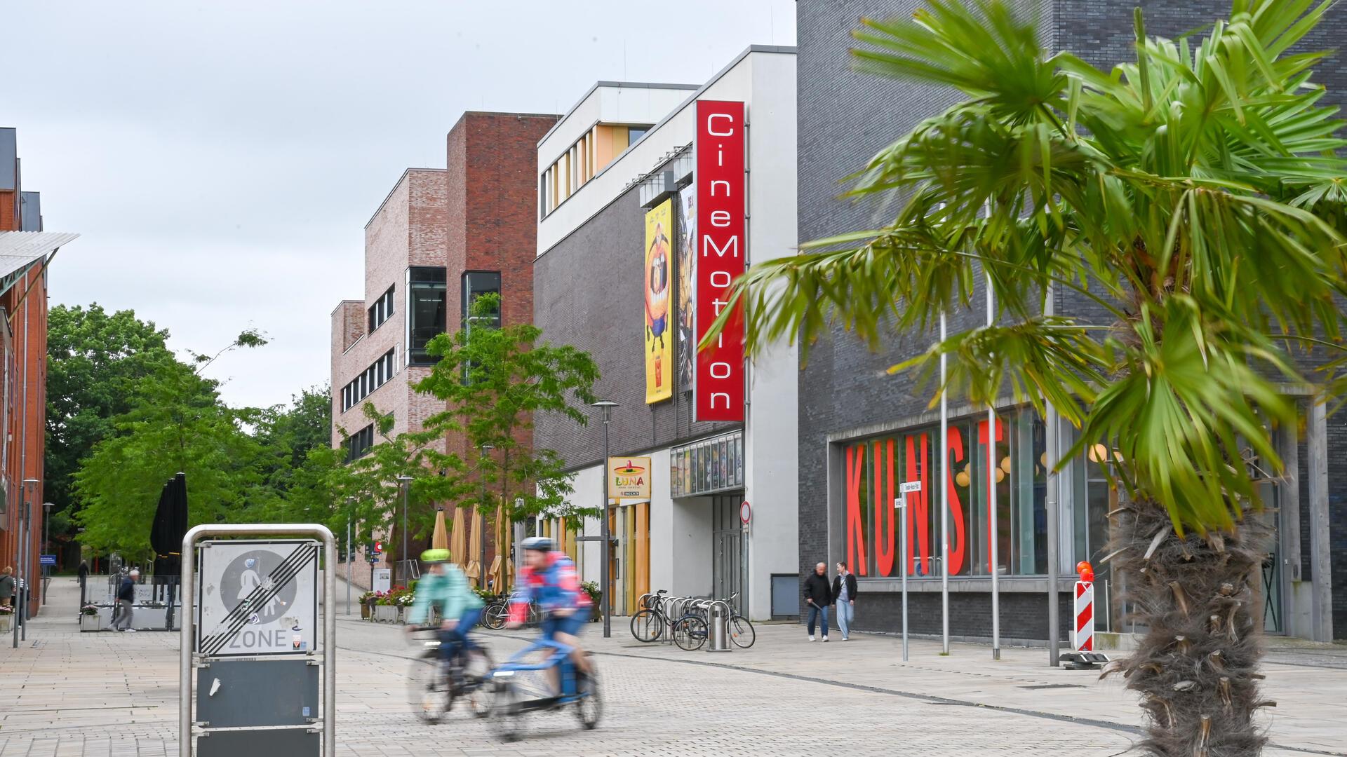 Blick auf das CineMotion-Kino in Bremerhaven mit rotem Fassadenschild. Im Vordergrund eine Palme und Radfahrende in Bewegung, rechts das Kunstmuseum mit großer „KUNST“-Beschriftung im Fenster.