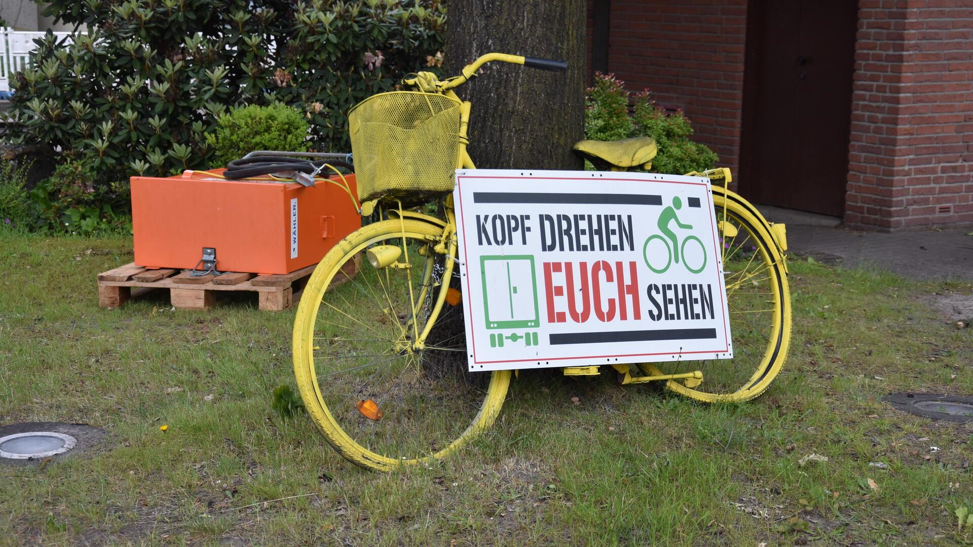 Ein gelbes Fahrrad mit dem Schild „Kopf drehen euch sehen“ weist an einer Kreuzung auf den Schulterblick hin.