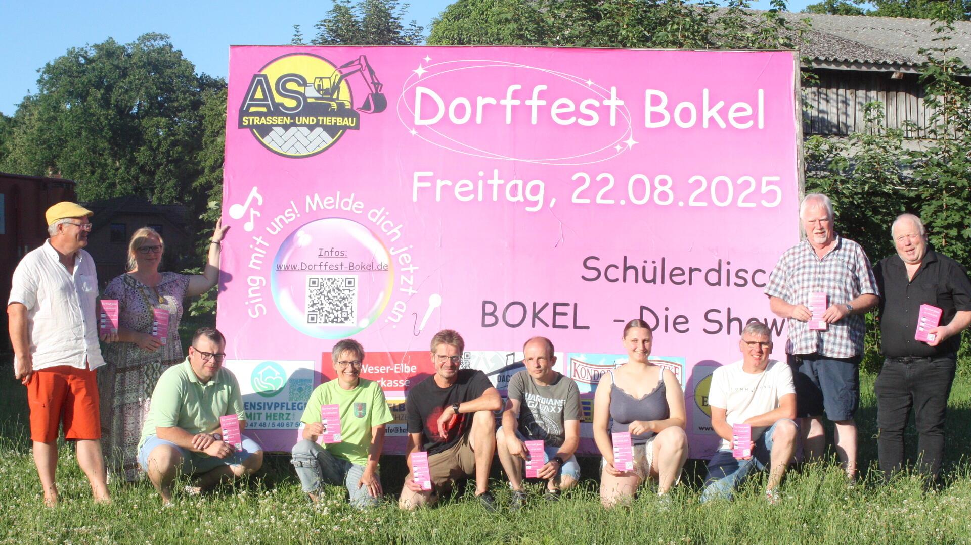 Mitglieder des Organisationsteams des Dorffests Bokel stehen und knien vor einem großen pinken Plakat mit Veranstaltungsinfos und QR-Code, aufgenommen bei Sonnenschein auf einer Wiese.