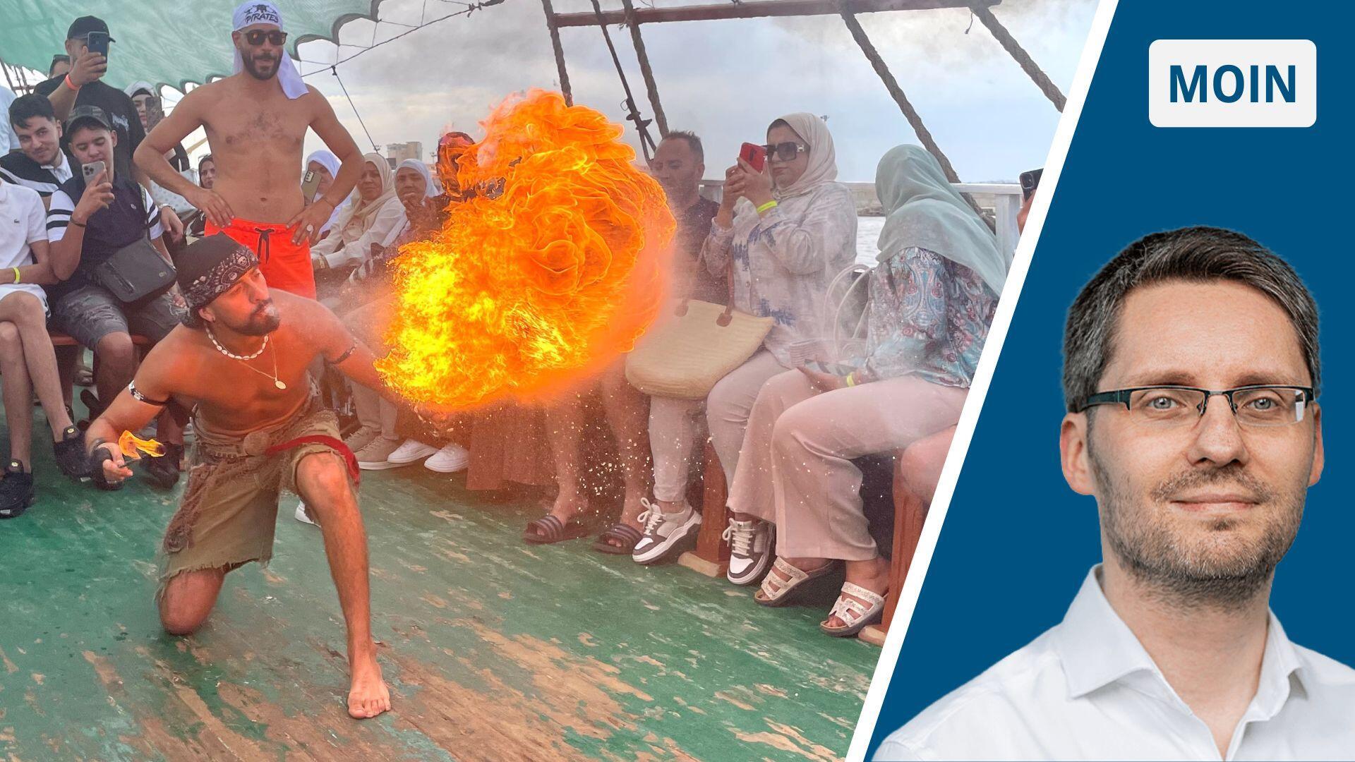Ein Mann, der auf dem Deck eines Schiffes kniet. Er pustet eine Flüssigkeit aus, mit der er einen Feuerball erzeugt. Publikum sitzt um ihn herum.