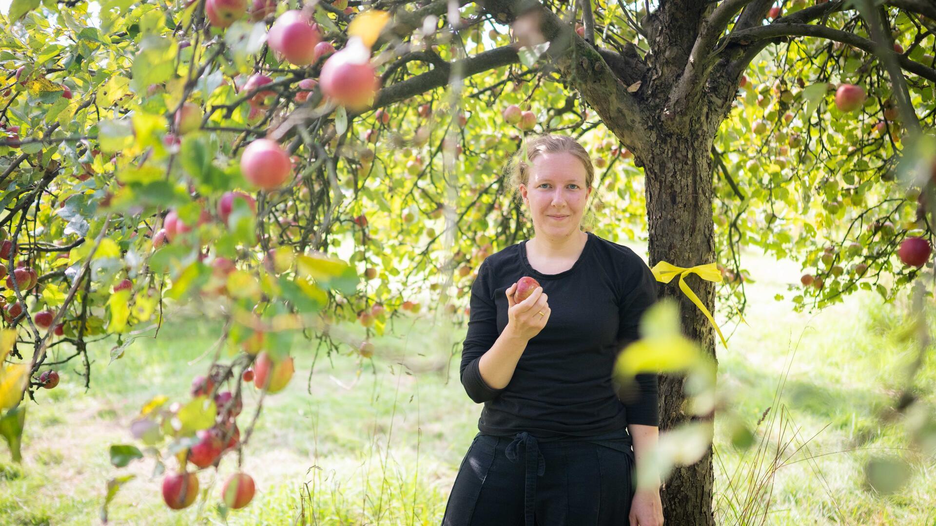 Eine Frau steht unter einem Baum und hält einen roten Apfel in der rechten Hand