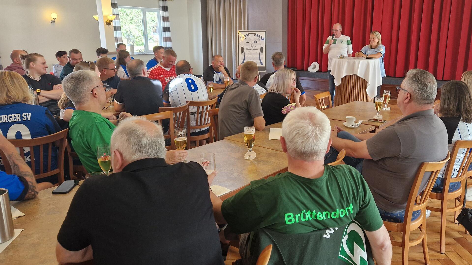 Auf einem Festsaal sitzen Menschen an langen Tischen. Vor der Bühne steht ein Mann mit einem Mikrofon in der Hand und eine Frau an einem Stehtisch.