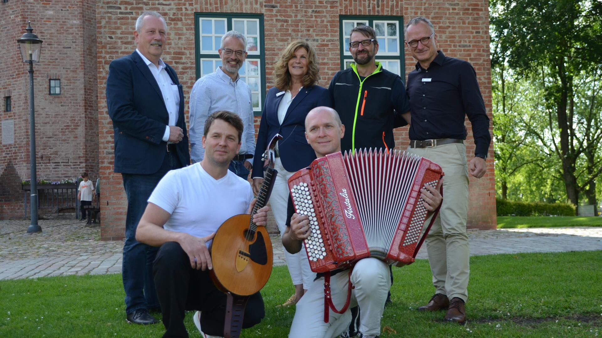 Sieben Personen posieren vor der Backsteinfassade der Burg Bederkesa. Zwei Musiker knien vorn mit Mandoline und Akkordeon, fünf weitere Personen stehen dahinter auf dem Rasen.