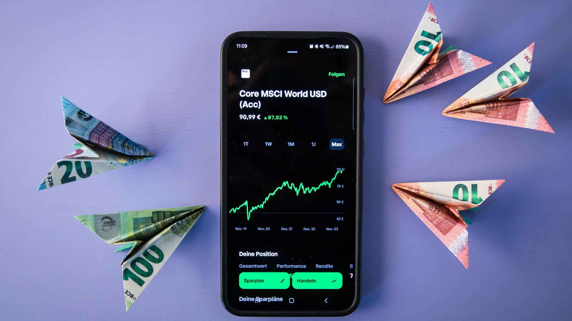 Ein Smartphone und Geldscheine liegen auf einem Tisch. Auf dem Smartphone wird der Kurs des MSCI World ETF angezeigt.