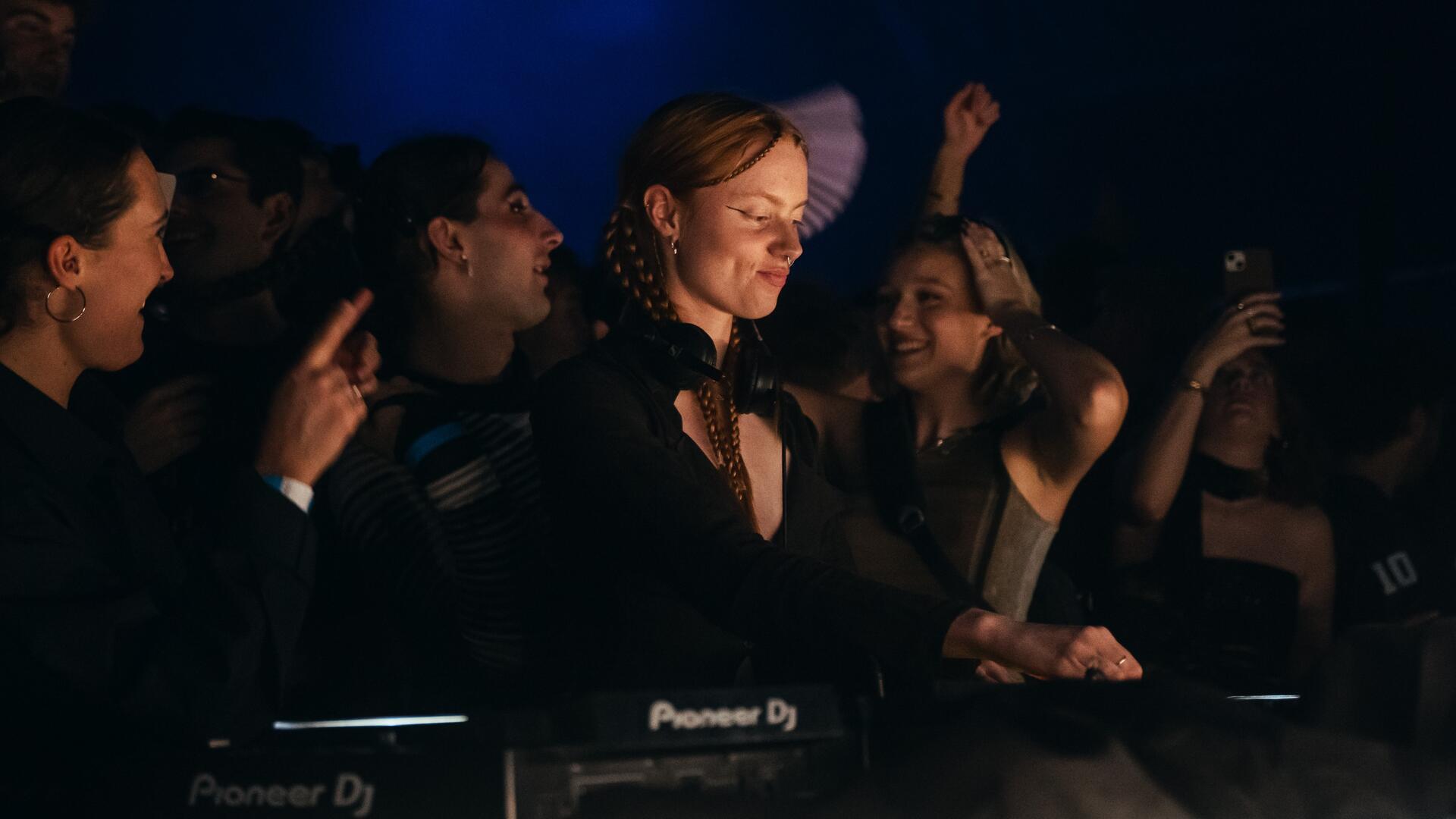 Eine junge Frau steht lächelnd an einem DJ-Pult mit Kopfhörern um den Hals. Um sie herum tanzen und jubeln mehrere Personen in einer dunklen, stimmungsvollen Club-Atmosphäre.