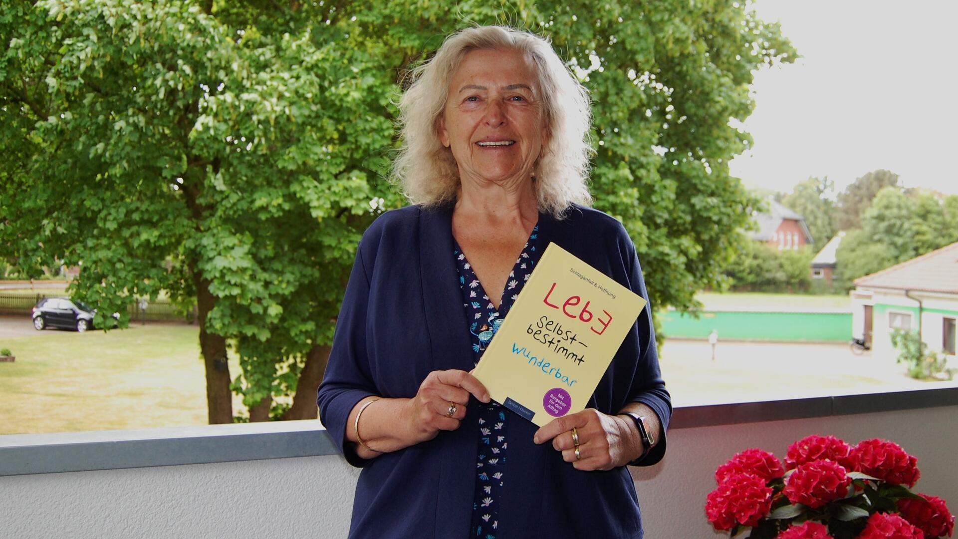 Eine ältere Frau mit blond-grauem Haar steht lächelnd auf einem Balkon vor grünem Baumbestand. Sie hält ein Buch mit dem Titel „Lebe selbstbestimmt wunderbar“ in den Händen. Auf dem Balkon stehen rote Blumen.
