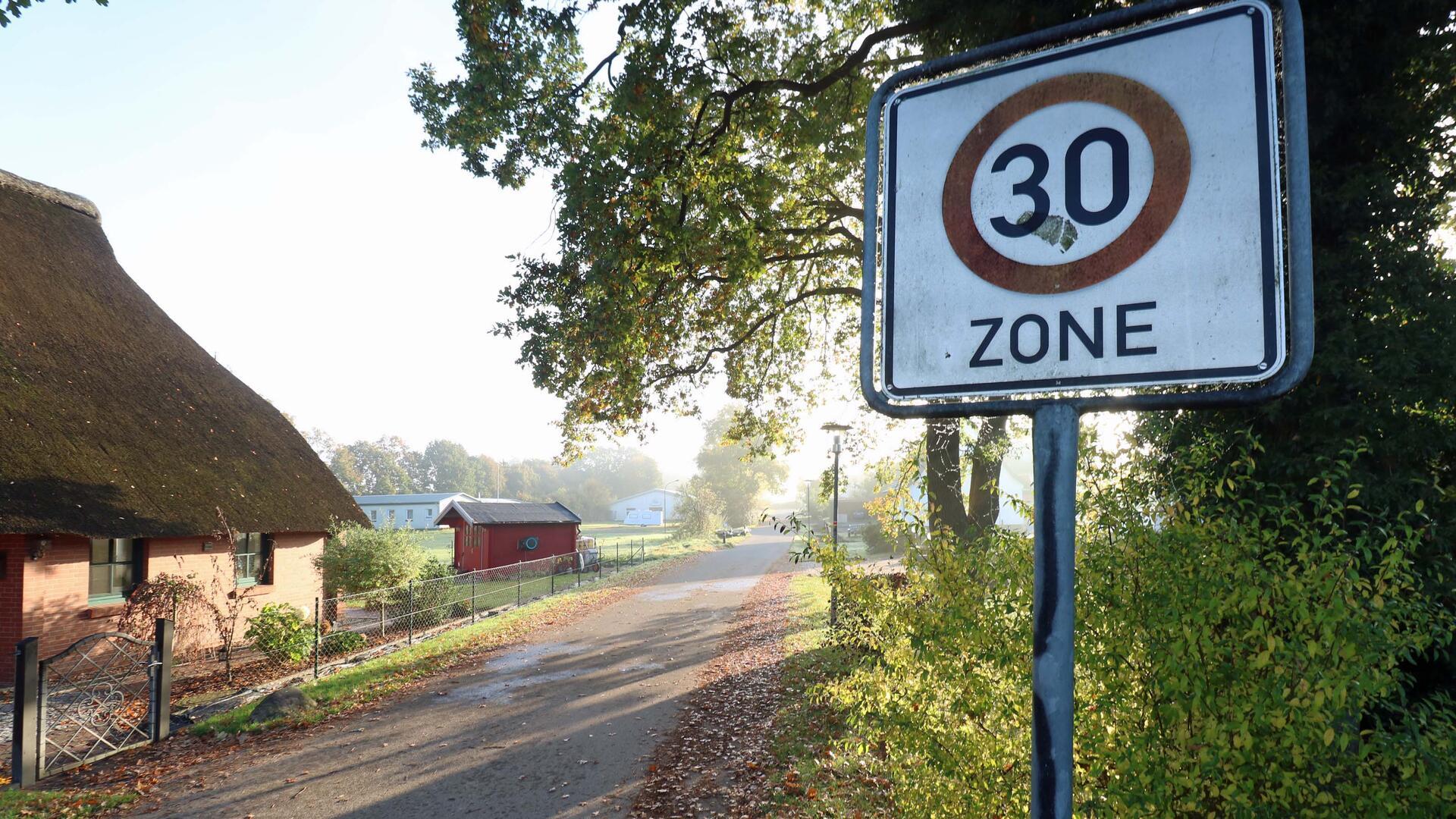 Eine Dorfstraße. Links ein Reetdachhaus, auf der rechten Straßenseite Gebüsch, ein Baum und ein Tempo-30-Schild