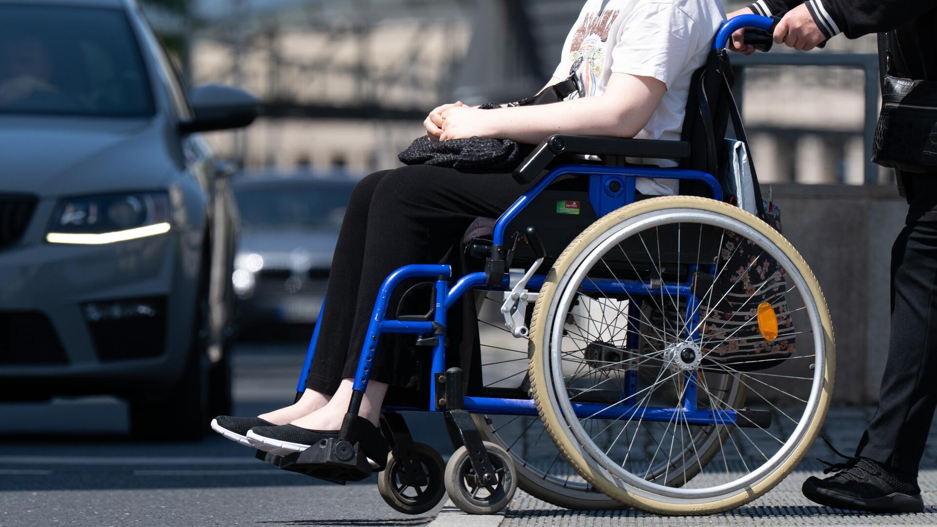 Person im Rollstuhl wird von einer Begleitperson auf einem Gehweg an einer Straße geschoben; im Hintergrund sind Autos im Stadtverkehr zu sehen.