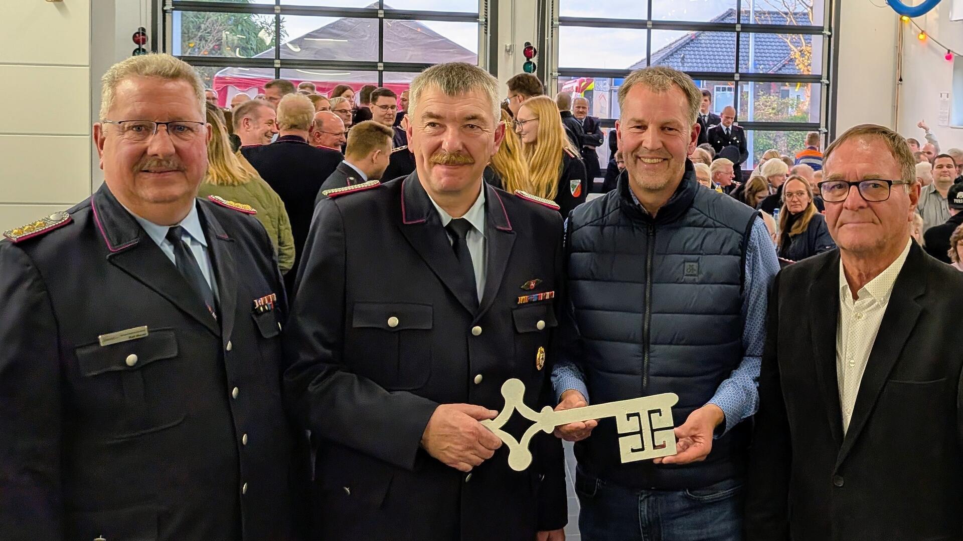 Vier Männer stehen bei der Einweihung eines Feuerwehrhauses zusammen, zwei von ihnen tragen Uniform. Einer hält einen symbolischen Schlüssel, im Hintergrund sind viele Gäste zu sehen.