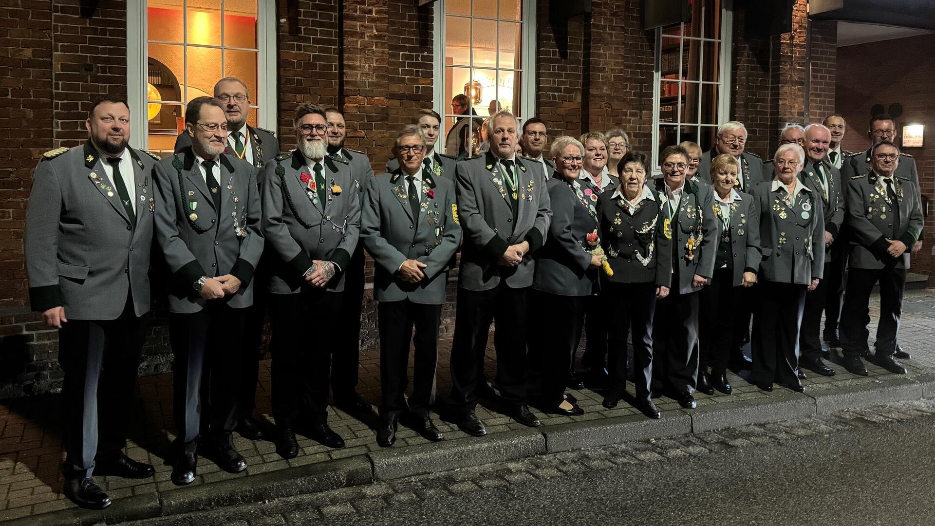 Eine Gruppe von Personen in grauen Uniformjacken mit Abzeichen steht in einer Reihe vor einem Backsteingebäude. Die Gruppe besteht aus Männern und Frauen unterschiedlichen Alters, die lächeln und für ein Foto posieren.