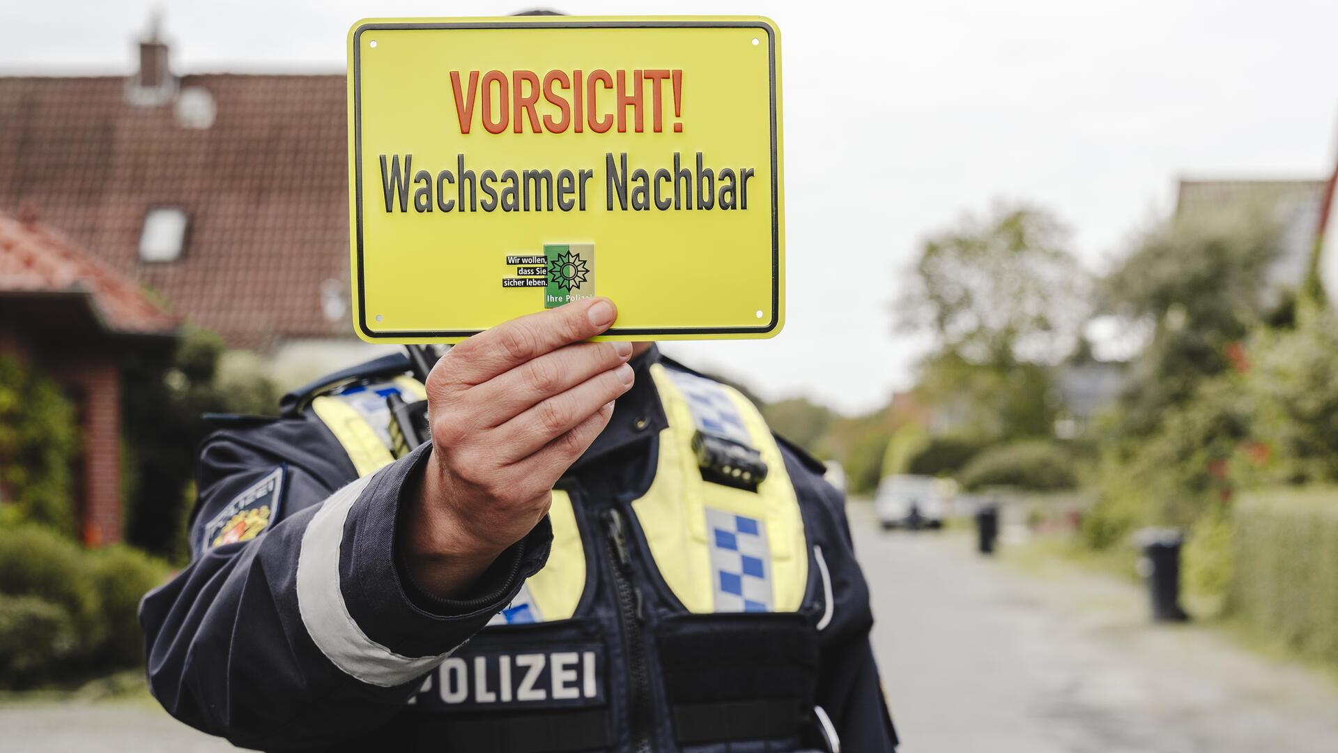 Ein Polizeibeamter hält ein gelbes Schild mit der Aufschrift „Vorsicht! Wachsamer Nachbar“ in die Kamera. Im Hintergrund ist eine ruhige Wohnstraße zu sehen.