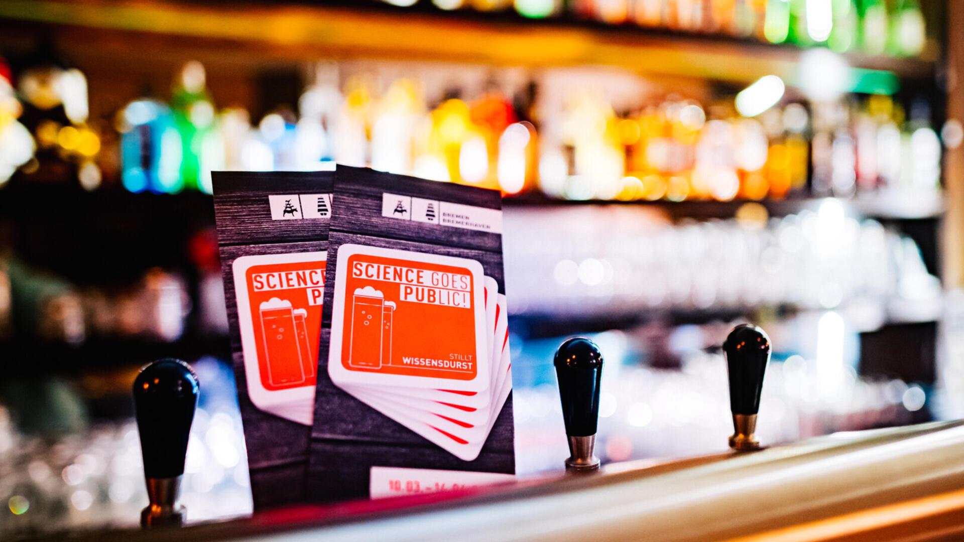 Zwei Flyer der Veranstaltungsreihe „Science goes Public!“ stehen auf einer Theke in einer Bar, im Hintergrund unscharf bunte Flaschen und Zapfhähne.