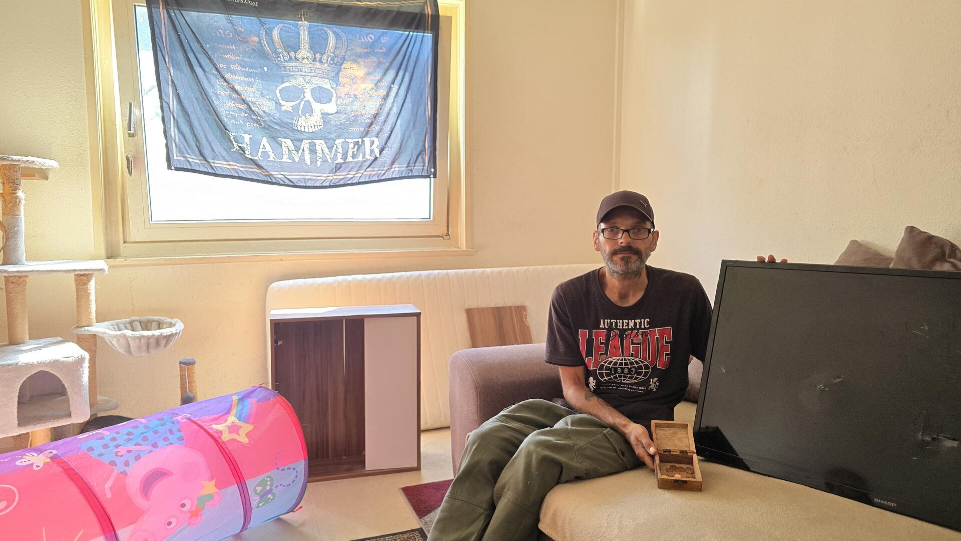 Ein Mann mit Kappe und Brille sitze auf einr Couch, er hält eine leere Holzschachtel und einen zerstörten Fernseher