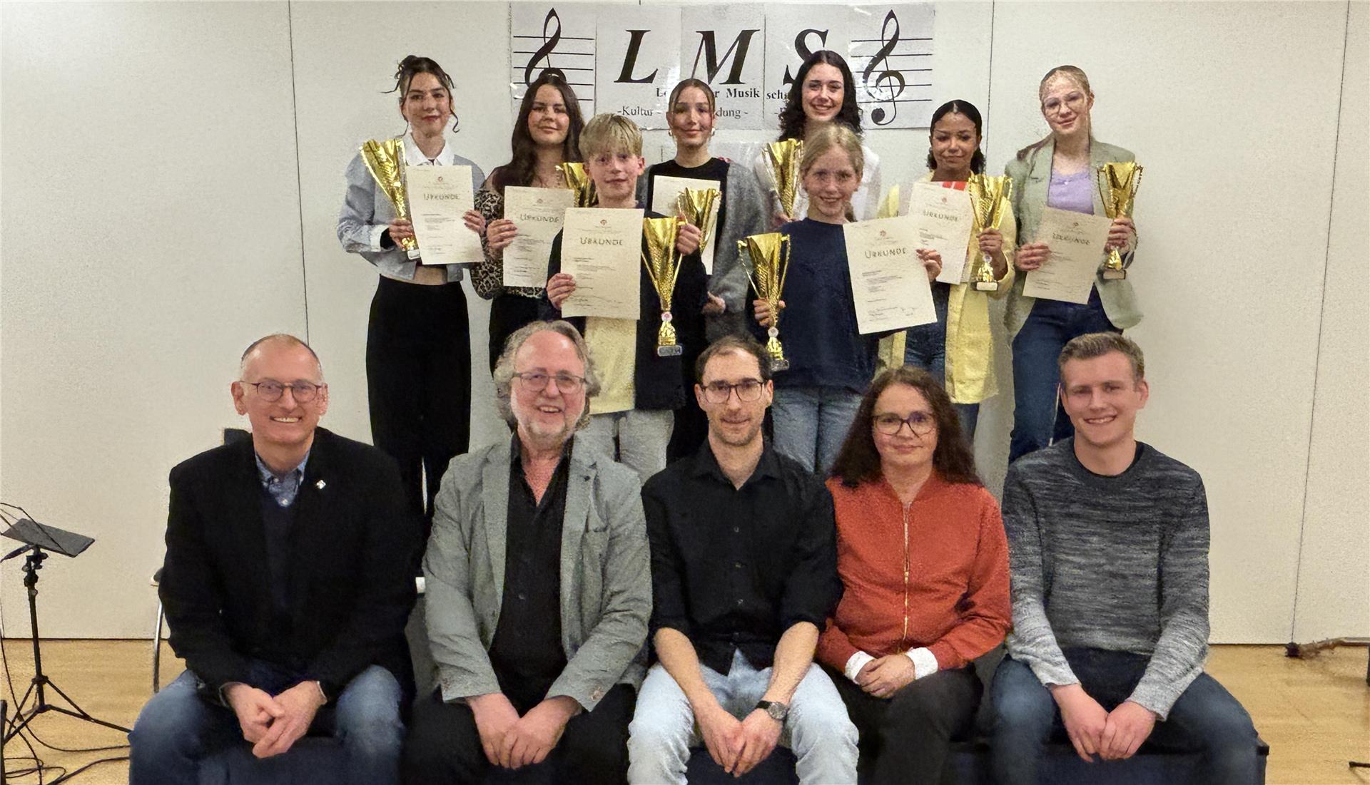 Acht junge Musikerinnen und Musiker stehen mit Urkunden und Pokalen in den Händen. Vor ihnen sitzen fünf Erwachsene. Im Hintergrund hängt das Logo der Loxstedter Musikschule.