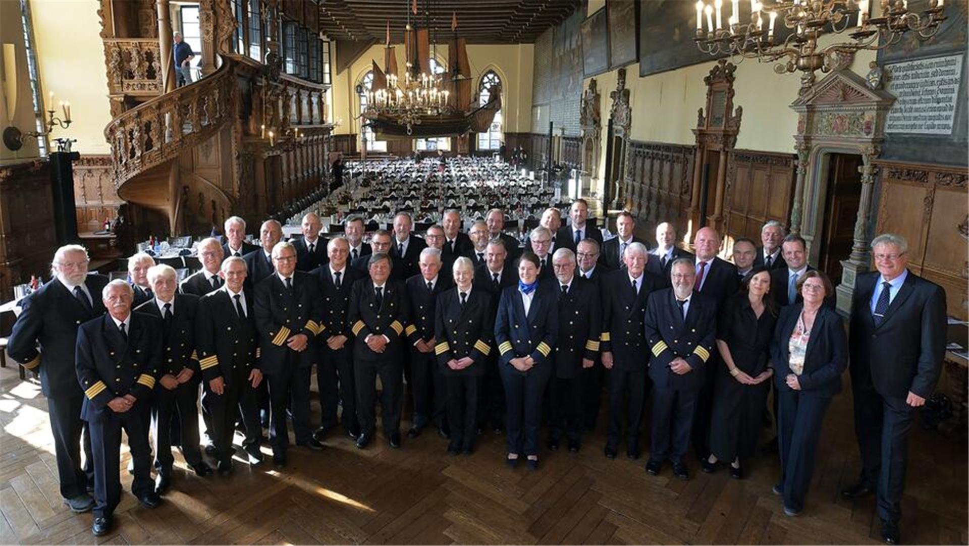 Gruppenfoto von Kapitänen und Gästen in Uniform und festlicher Kleidung beim 58. Bremer Kapitänstag in der historischen Oberen Rathaushalle mit prunkvollem Holzinterieur und großer Schiffsreplik an der Decke. Im Hintergrund sind festlich gedeckte Tische zu sehen.
