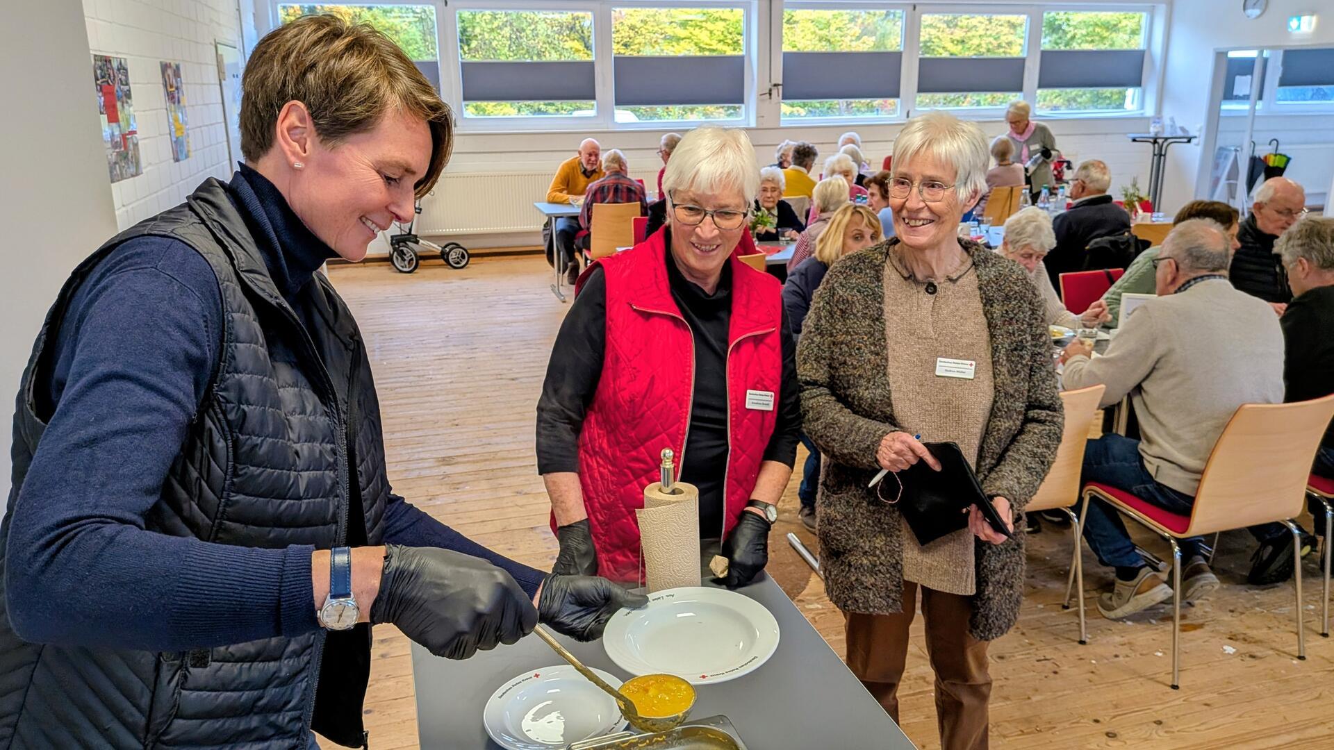 Menschen sitzen in einem Raum an Tischen. Im Vordergrund wird von einer Frau mit einer Kelle Suppe auf einen Teller gelöffelt.