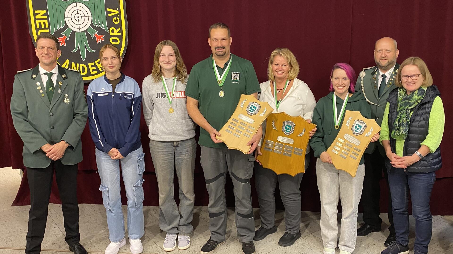 Auf dem Foto stehen acht Personen nebeneinander vor einer weinroten Bühne mit dem Vereinswappen des Schützenvereins Neuenlandermoor. Mehrere tragen grüne Schützenuniformen oder Vereinskleidung, einige haben Medaillen um den Hals. Drei Personen in der Mitte halten große Holztafeln mit eingravierten Namensschildern, die als Siegerplaketten dienen. Alle blicken in die Kamera und lächeln.