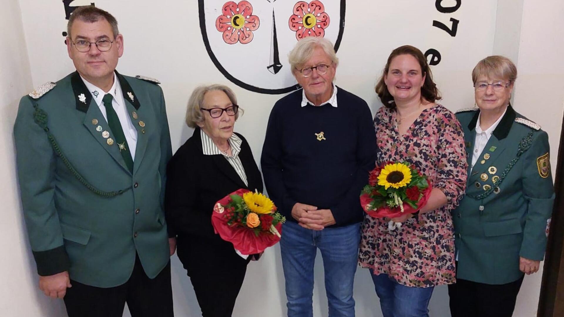 Fünf Personen stehen vor einem Wandwappen, zwei Männer und drei Frauen. Zwei Frauen und ein Mann in der Mitte halten Blumensträuße, die äußeren Personen tragen grüne Schützenuniformen mit Abzeichen.