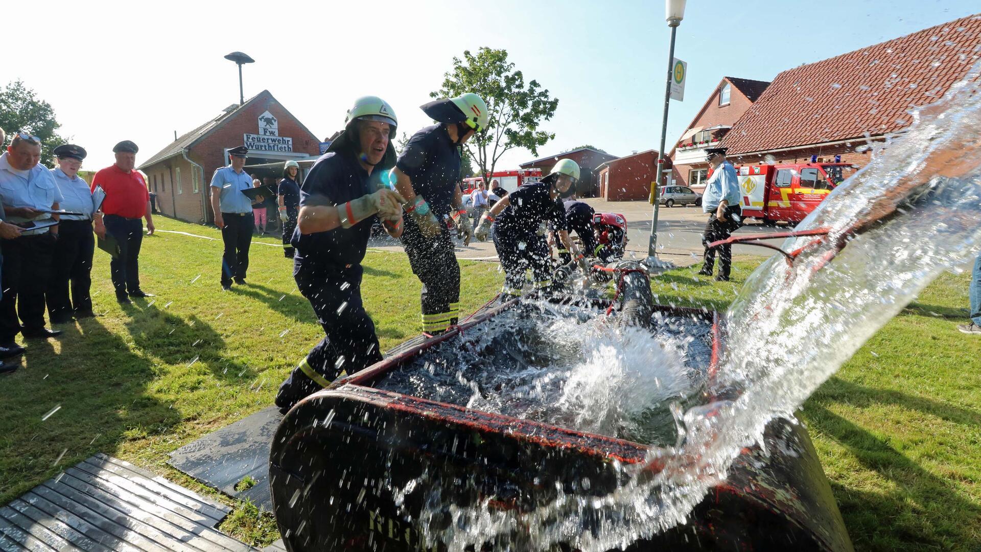 Feuerwehrleute in Einsatzkleidung pumpen Wasser aus einem Becken beim Fire Cup in Wurthfleth, während Juroren am Rand zuschauen und Feuerwehrfahrzeuge im Hintergrund stehen.