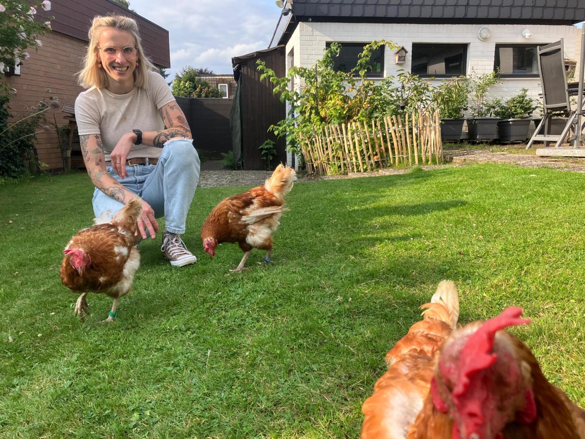 Eine blonde Frau hockt in ihrem Garten mit drei Hühnern.