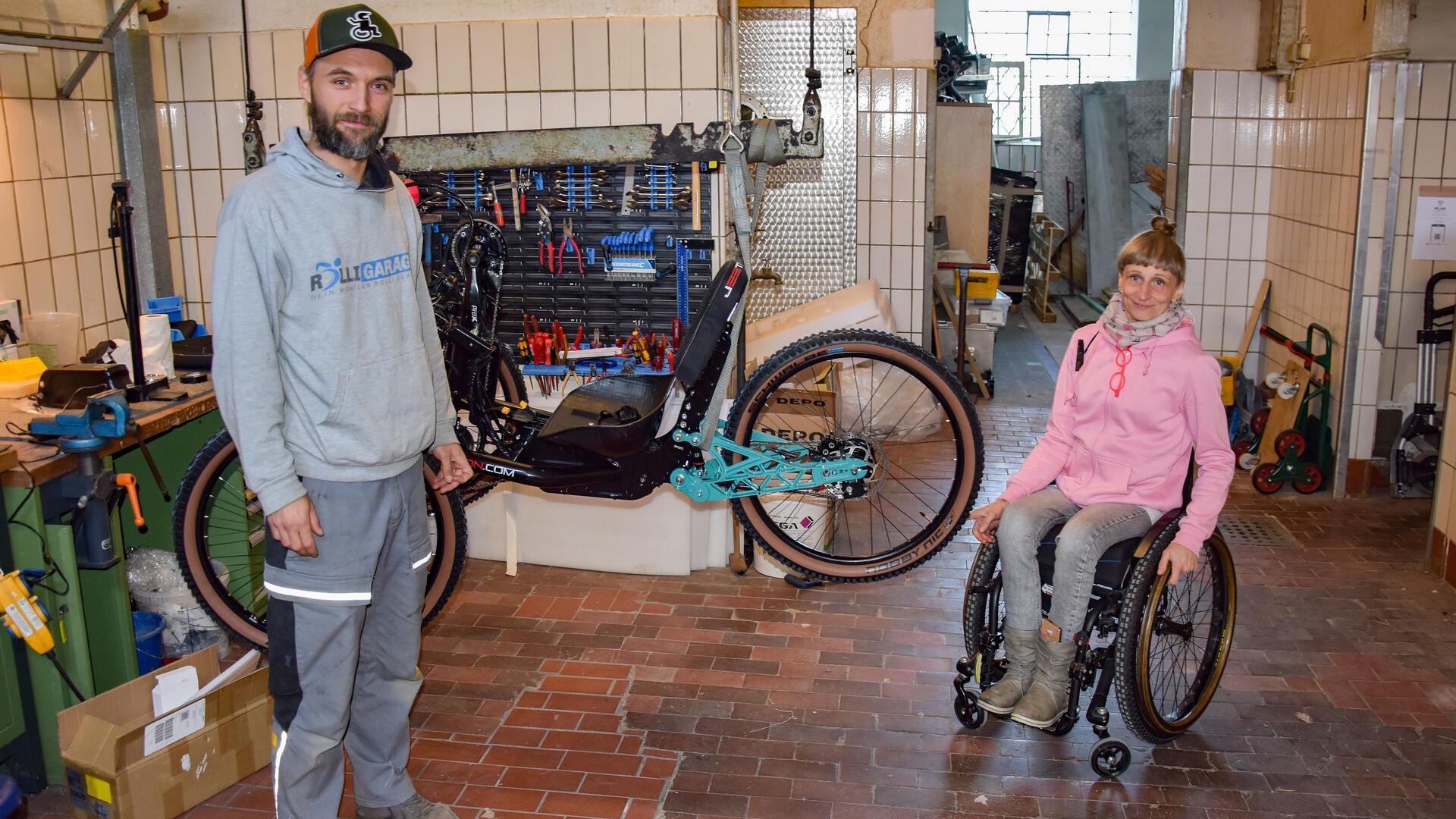 Ein Mann steht in einer Werkstatt, hinter ihm hängt ein Handbike am Haken, neben ihm sitzt eine Rollstuhlfahrerin.