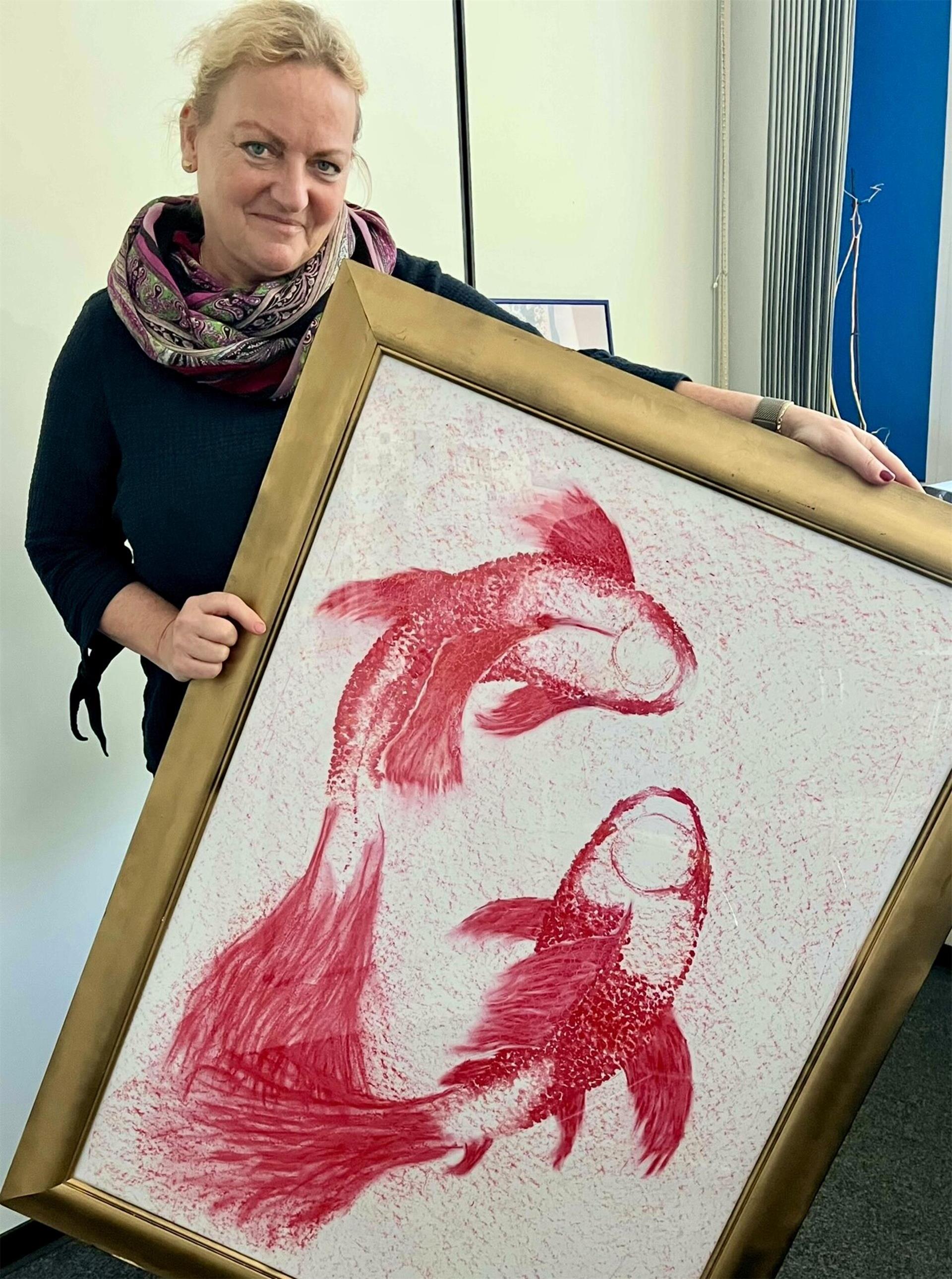 Eine Frau hält ein gerahmtes Kunstwerk mit zwei roten Fischen auf weißem Hintergrund. Sie lächelt leicht und steht vor einer hellen Wand mit Vorhängen im Hintergrund.