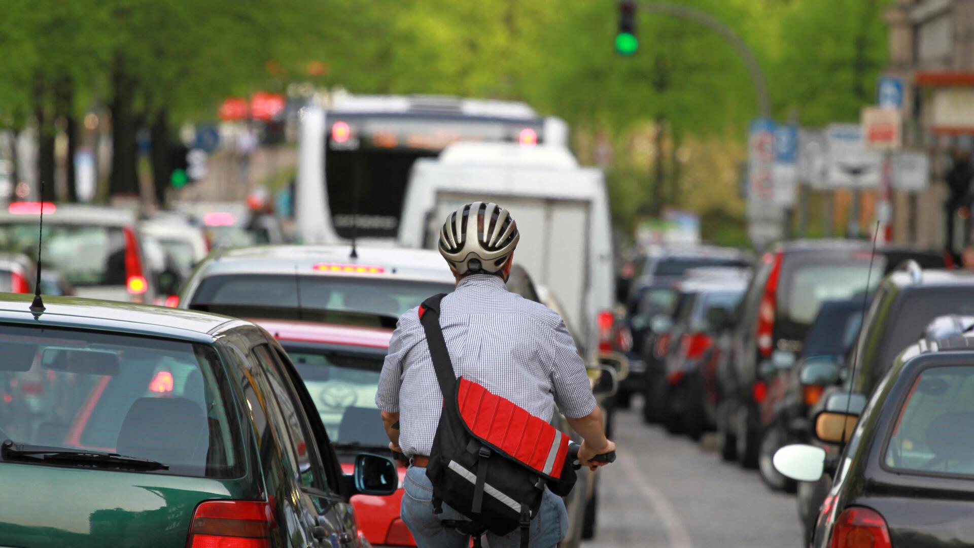 **ALT-Text:**
Ein Radfahrer mit Helm fährt zwischen dichtem Autoverkehr in einer städtischen Straße, umgeben von Autos, Bussen und Ampeln bei grünem Licht.