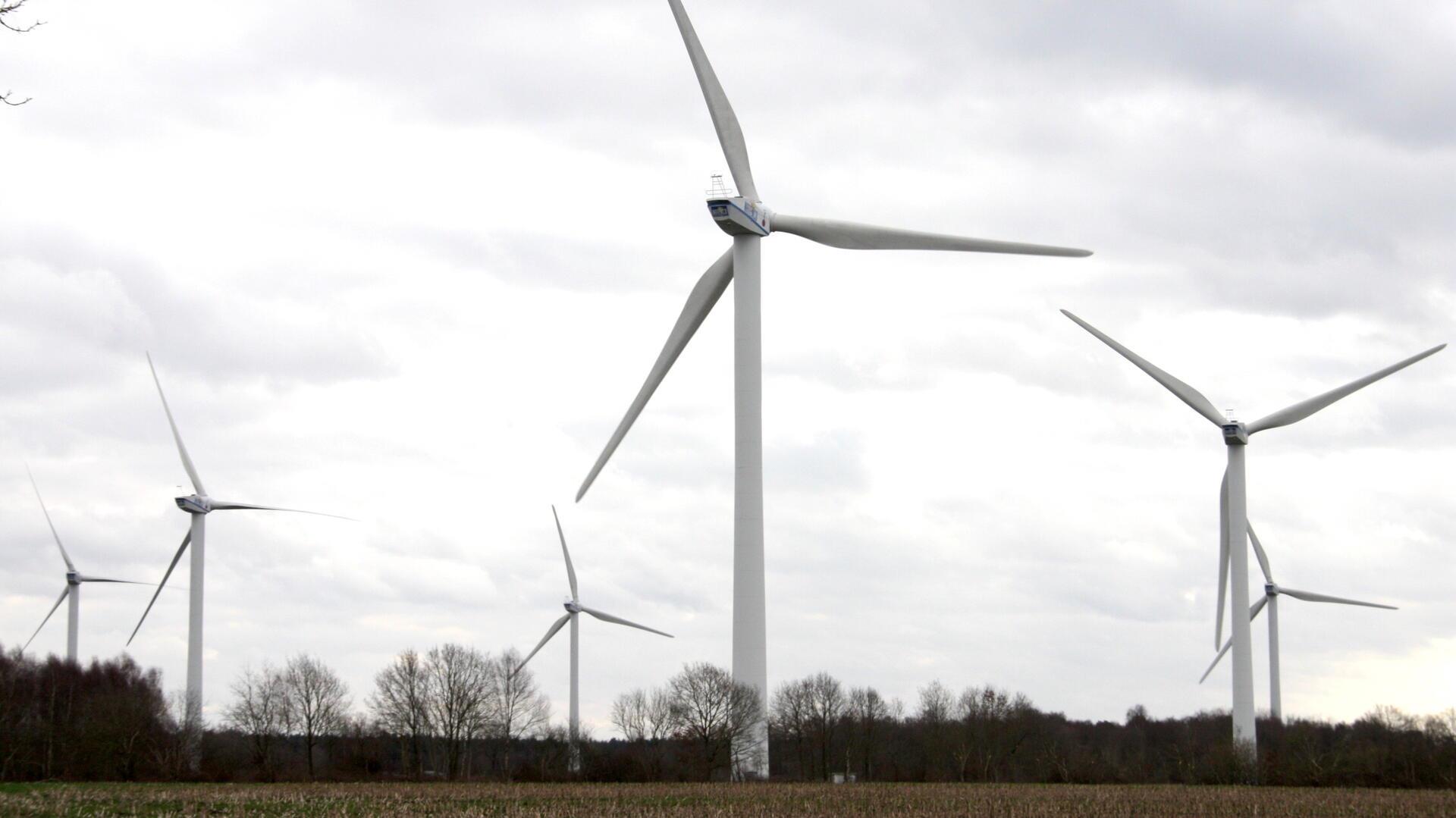 Mehrere große Windkraftanlagen stehen auf einem offenen Feld vor einem bewölkten Himmel. Im Hintergrund sind Bäume ohne Laub zu sehen. Die Szene wirkt ruhig und zeigt eine typische Landschaft mit moderner Windenergie-Nutzung.