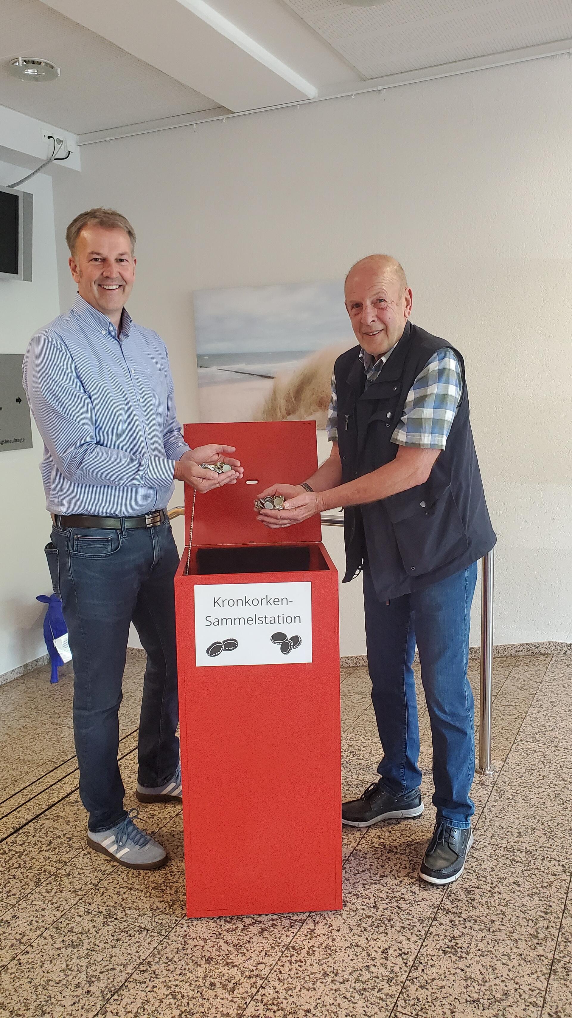 Beverstedts Gemeindebürgermeister Guido Dieckmann und Udo Kawaletz füllen die ersten Kronkorken in die neue Sammelstation im Beverstedter Rathaus.